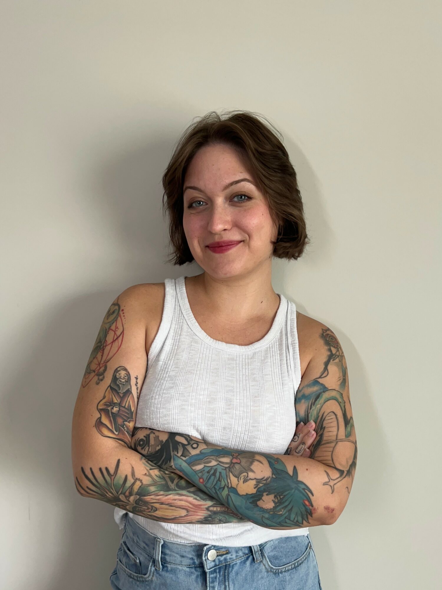 Young woman with short brown hair, wearing a white sleeveless top and jeans, standing with arms crossed against a plain wall.