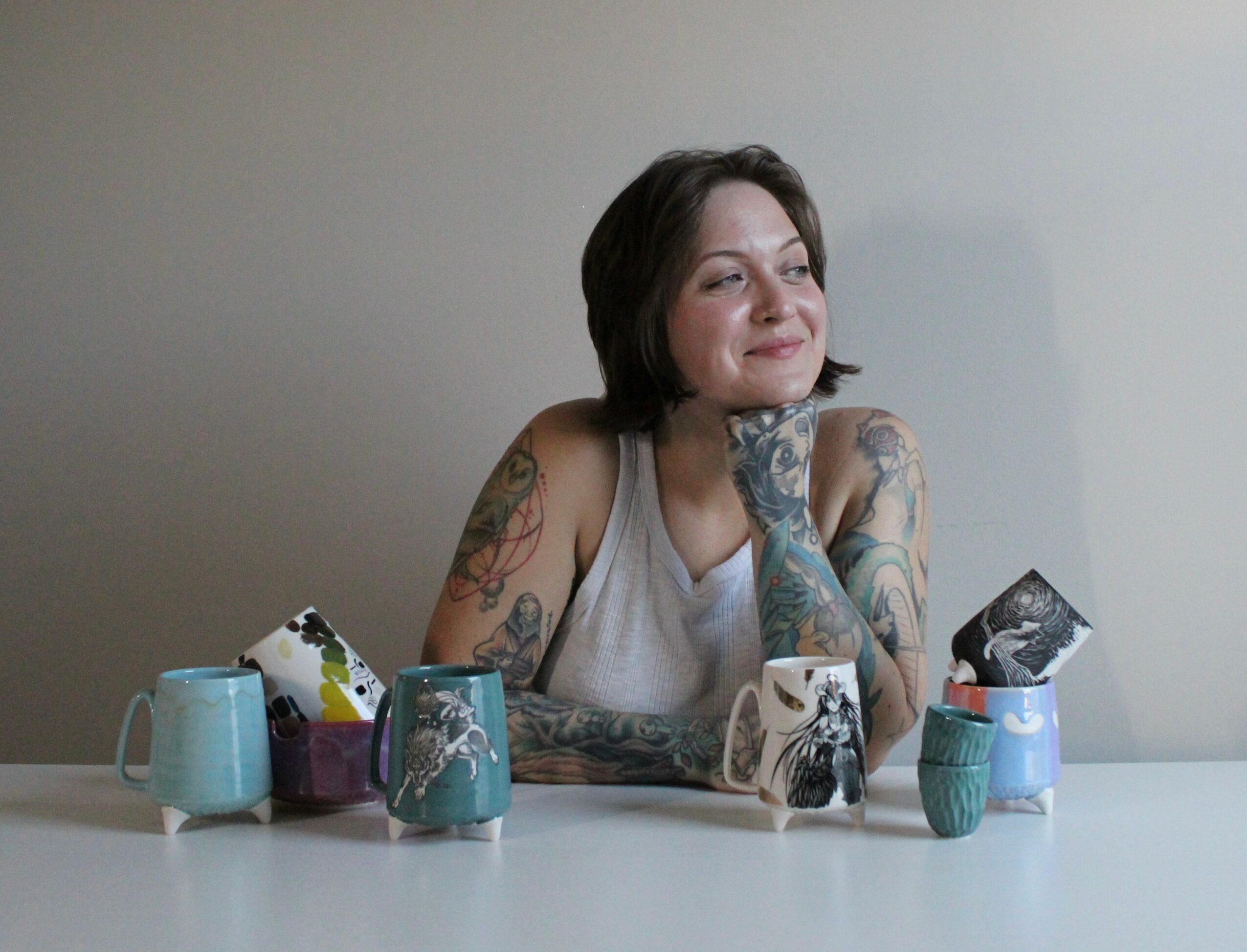 Woman with tattoos sitting at a table with colorful mugs, looking to the side, against a plain wall.