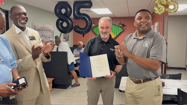 Three men standing indoors, one holding a certificate, celebrating an 85th birthday with balloons and decorations.