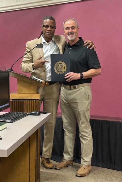 Two men standing together, one holding a certificate or award, in front of a pink wall.