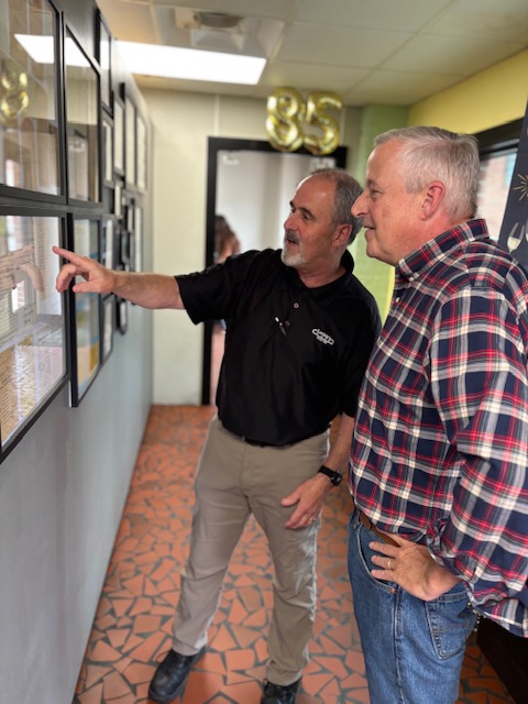 Two men look at framed pictures on a wall, one pointing, in a room with yellow walls and patterned floor.
