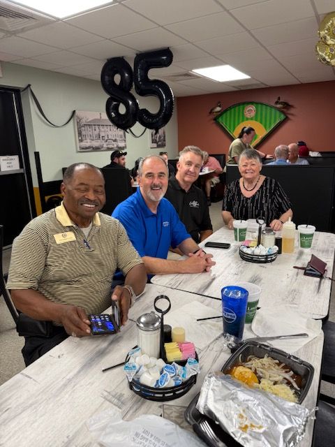 Four people gathered around a table with food and drinks, smiling and engaging in conversation in an office setting.