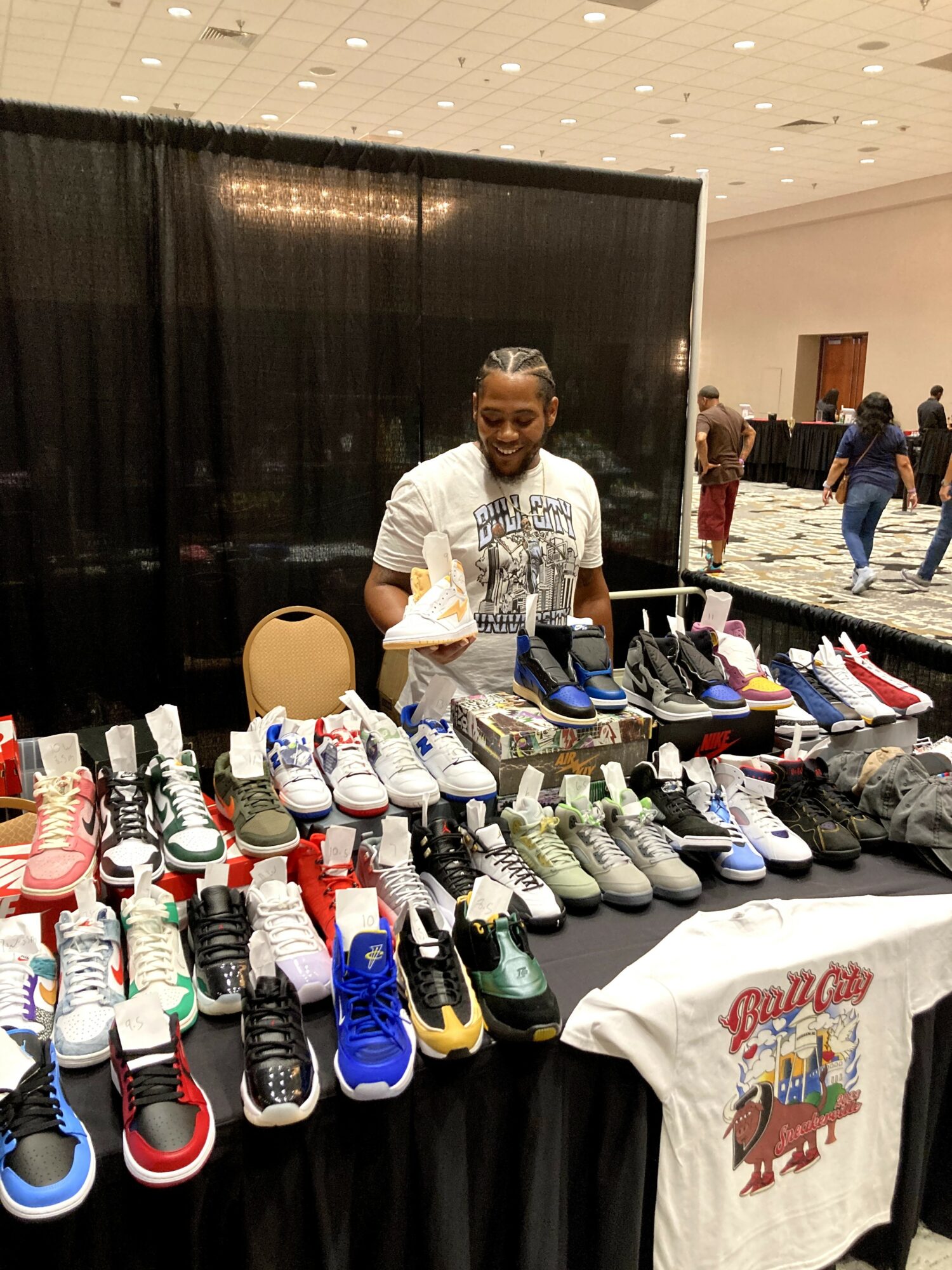 Woman standing behind a table with various sneakers and shoes at an indoor event, black curtain backdrop, people in background.