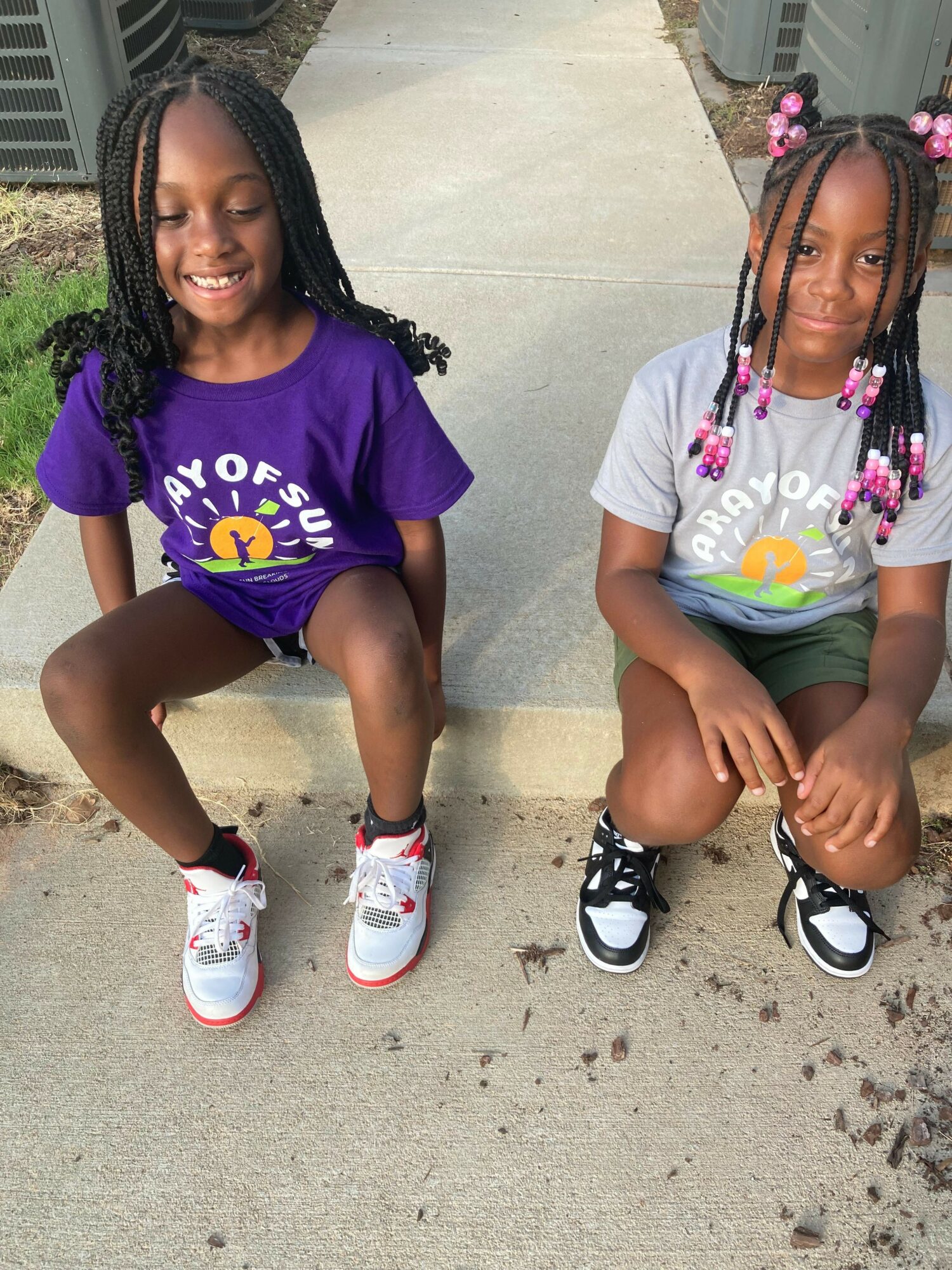 Two children sitting on the sidewalk, one with braided hair and colorful beads, the other with long black hair, both smiling.