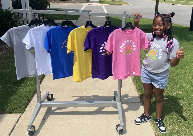 Young girl with braided hair and a gray T-shirt smiling next to a clothing rack with colorful T-shirts outdoors.