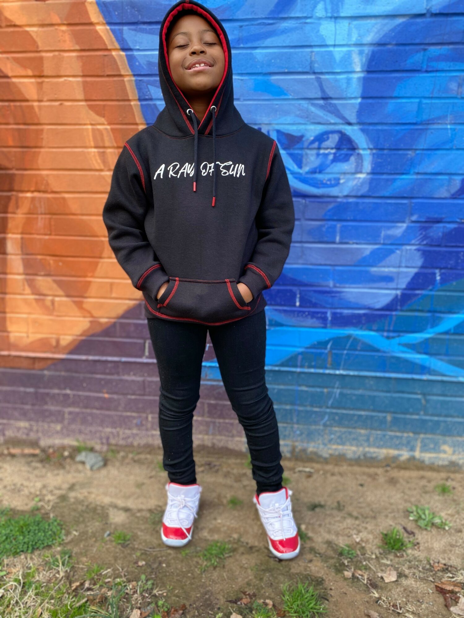 Child standing outdoors in front of a colorful mural, wearing a hoodie, black pants, and sneakers, smiling with eyes closed.