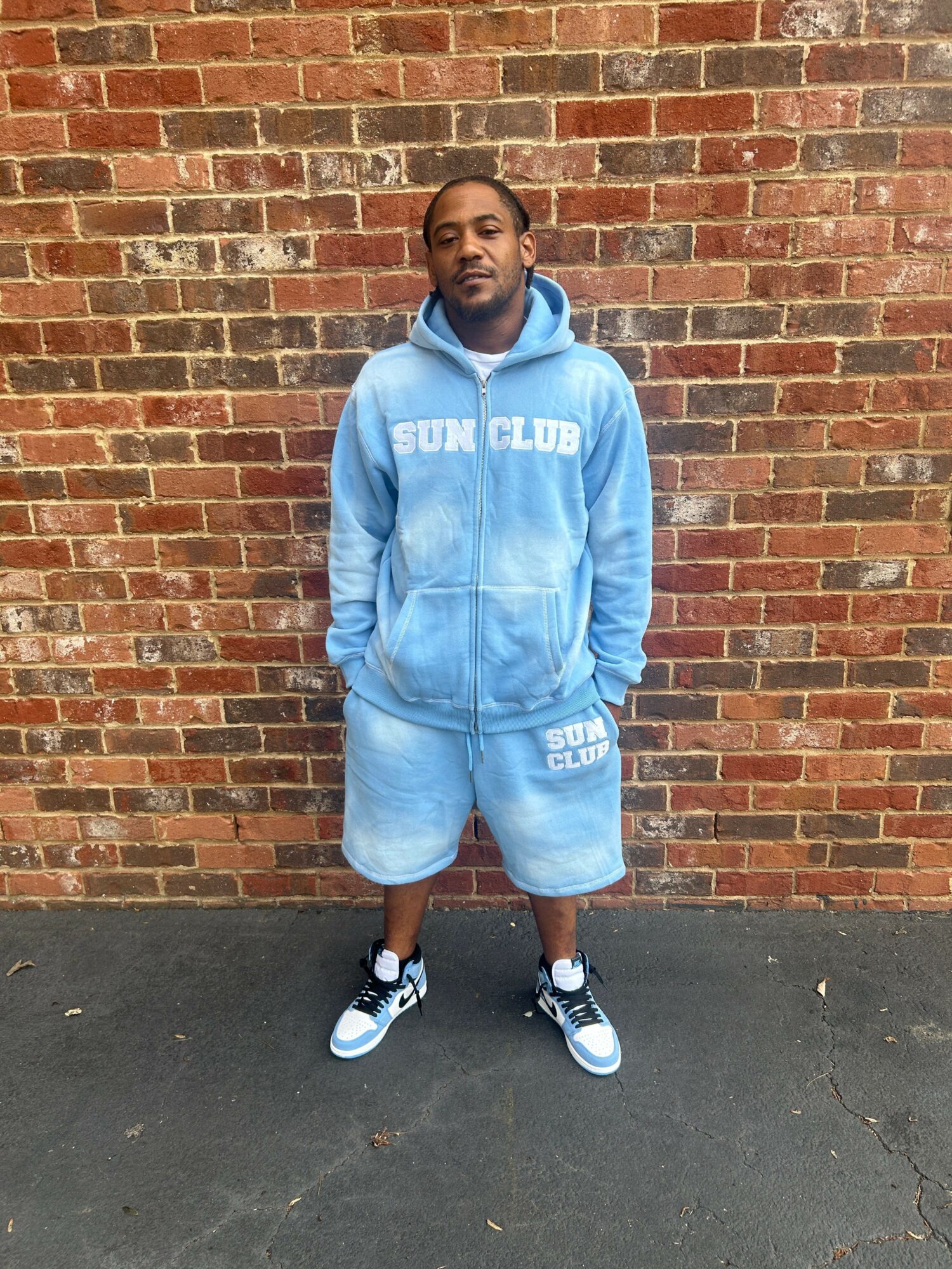 Young man in a light blue hoodie and shorts standing against a brick wall, facing forward, on a paved surface.