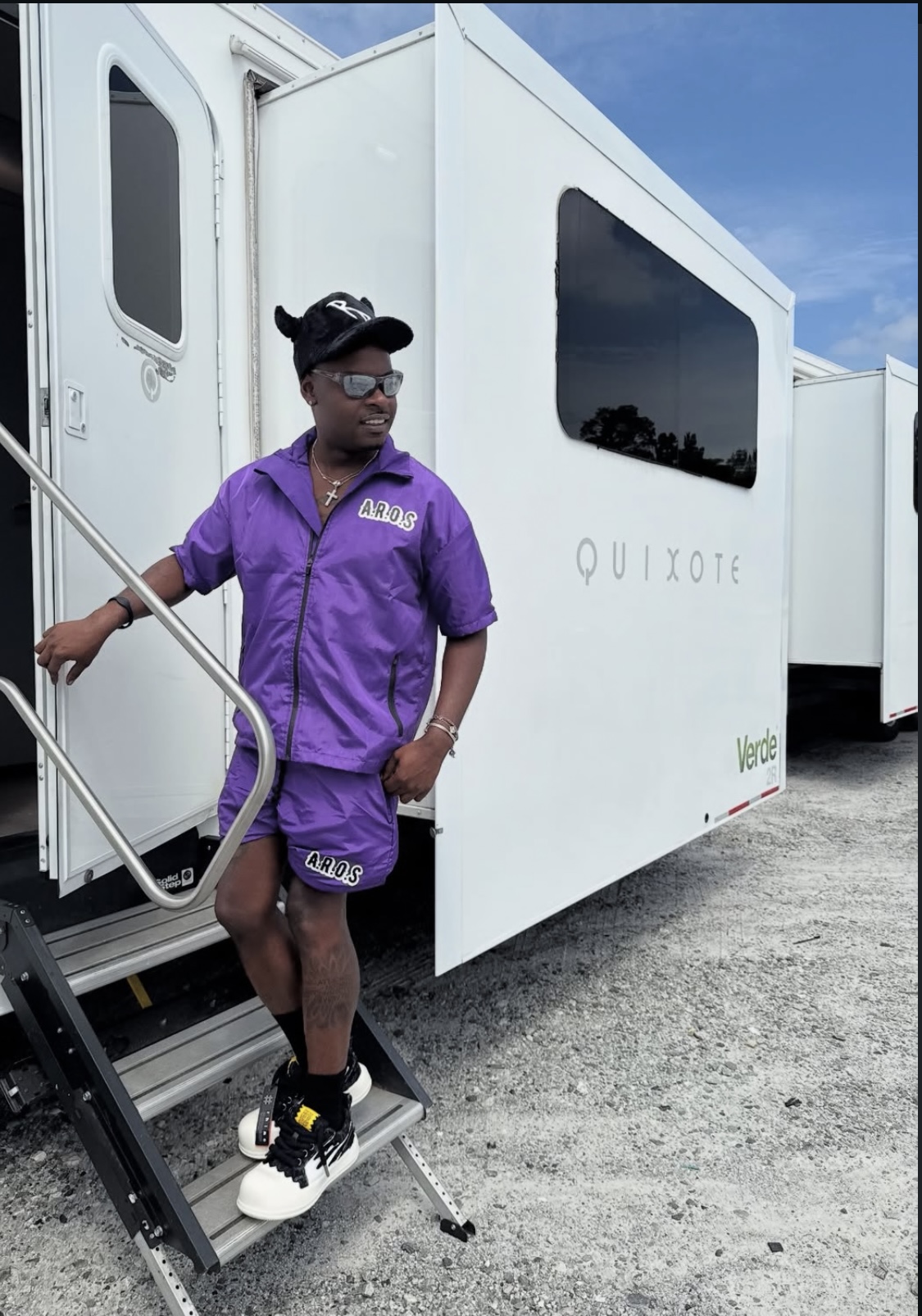 Person in purple outfit standing outside a white trailer with a blue sky background.
