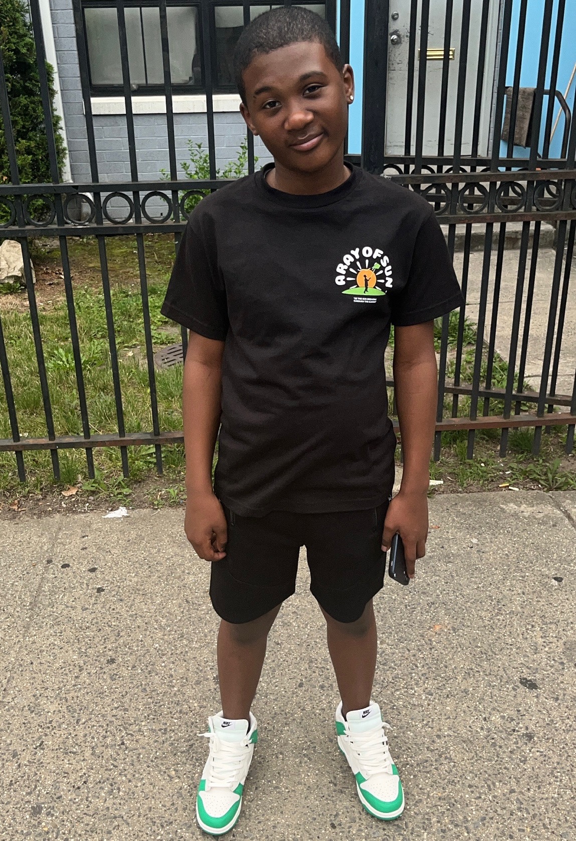 Young boy standing outdoors in front of a black fence, wearing a black t-shirt, black shorts, and white sneakers with green accents.