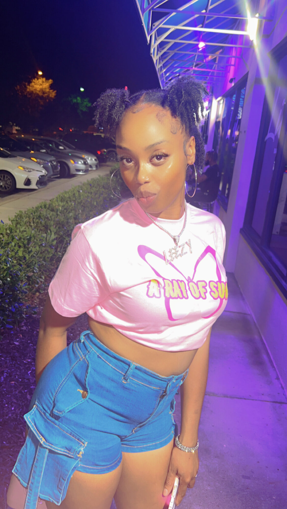 Young woman with styled hair, wearing a pink crop top and blue shorts, standing outdoors at night with colorful lights.