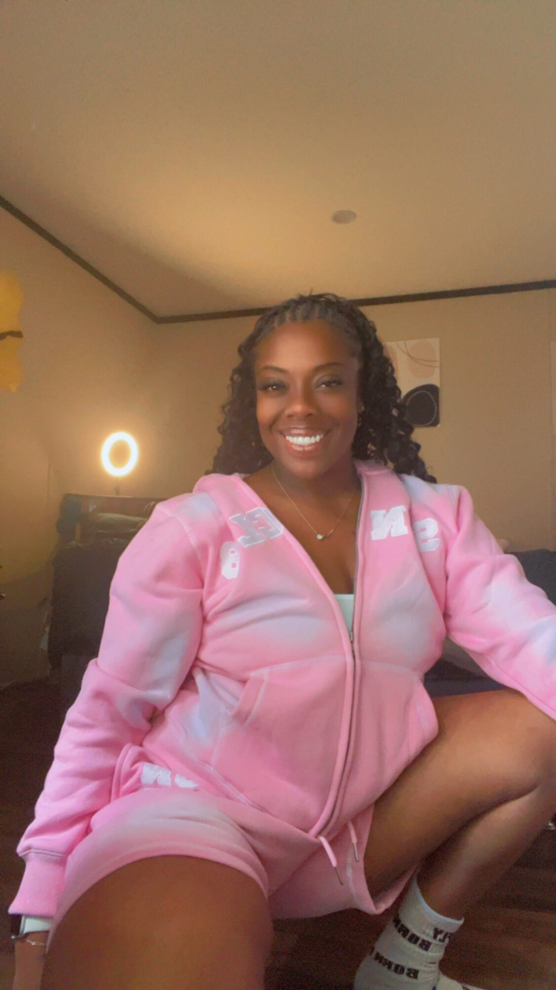 Smiling woman in pink hoodie and shorts sitting on the floor in a room with warm lighting.