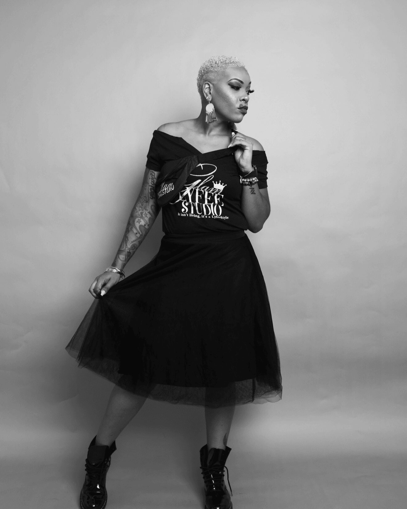 Woman with short platinum hair, tattoos, wearing a black off-shoulder dress and boots, standing against a plain background.