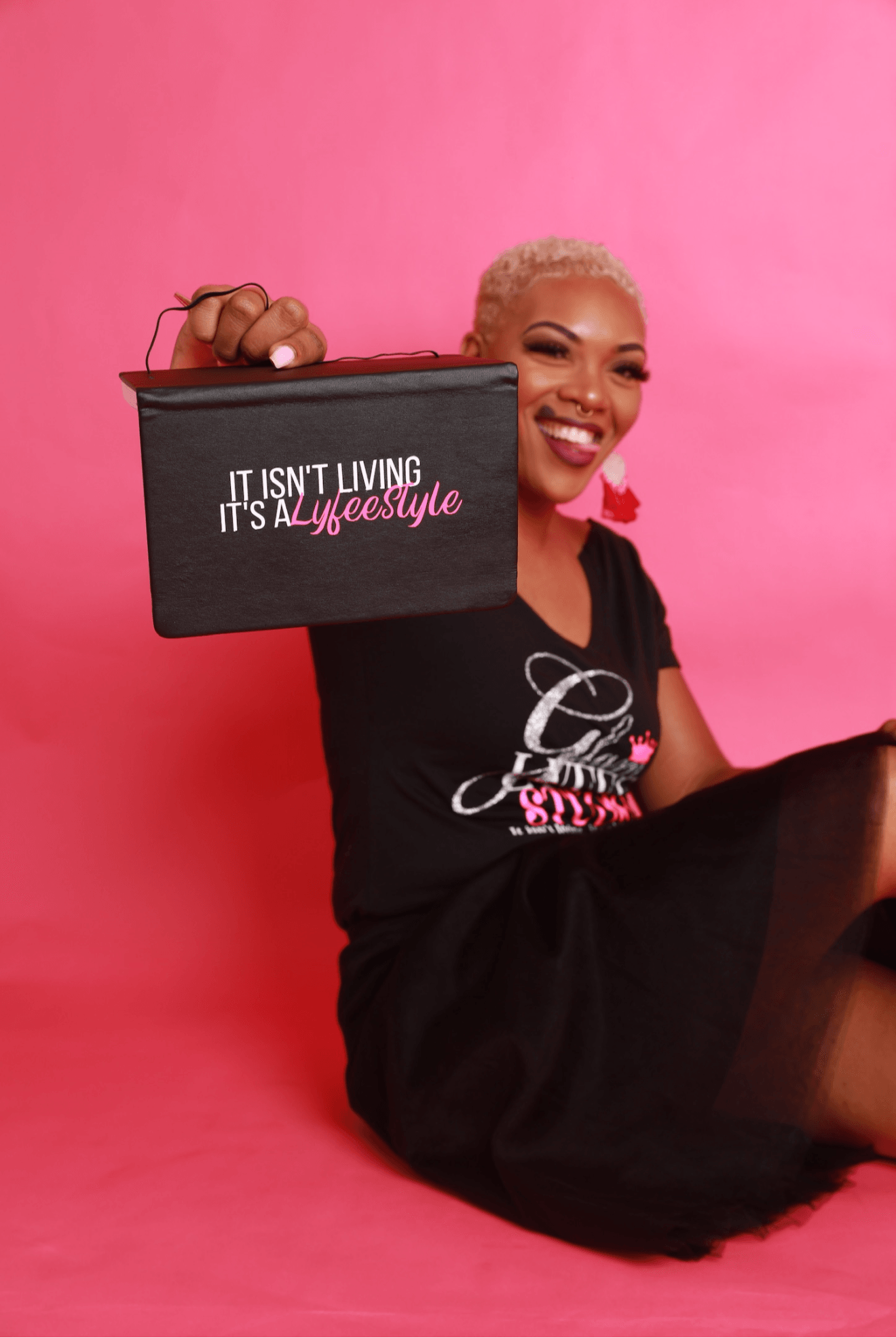 Smiling woman with short blonde hair holding a black bag with pink and white text, sitting against a pink background.