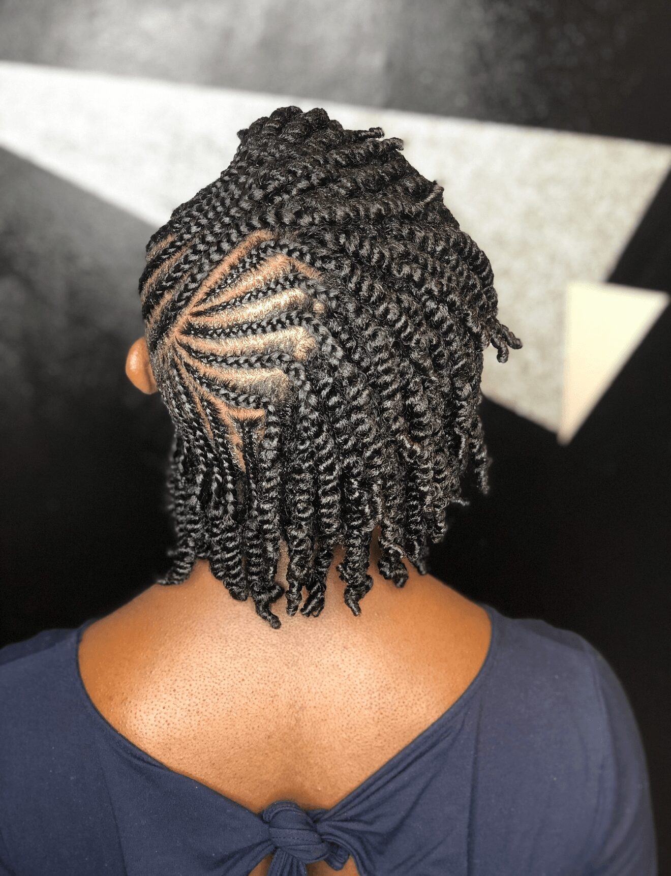 Back view of a person with short, twisted hairstyle and intricate scalp braids, wearing a dark top with a bow at the back.