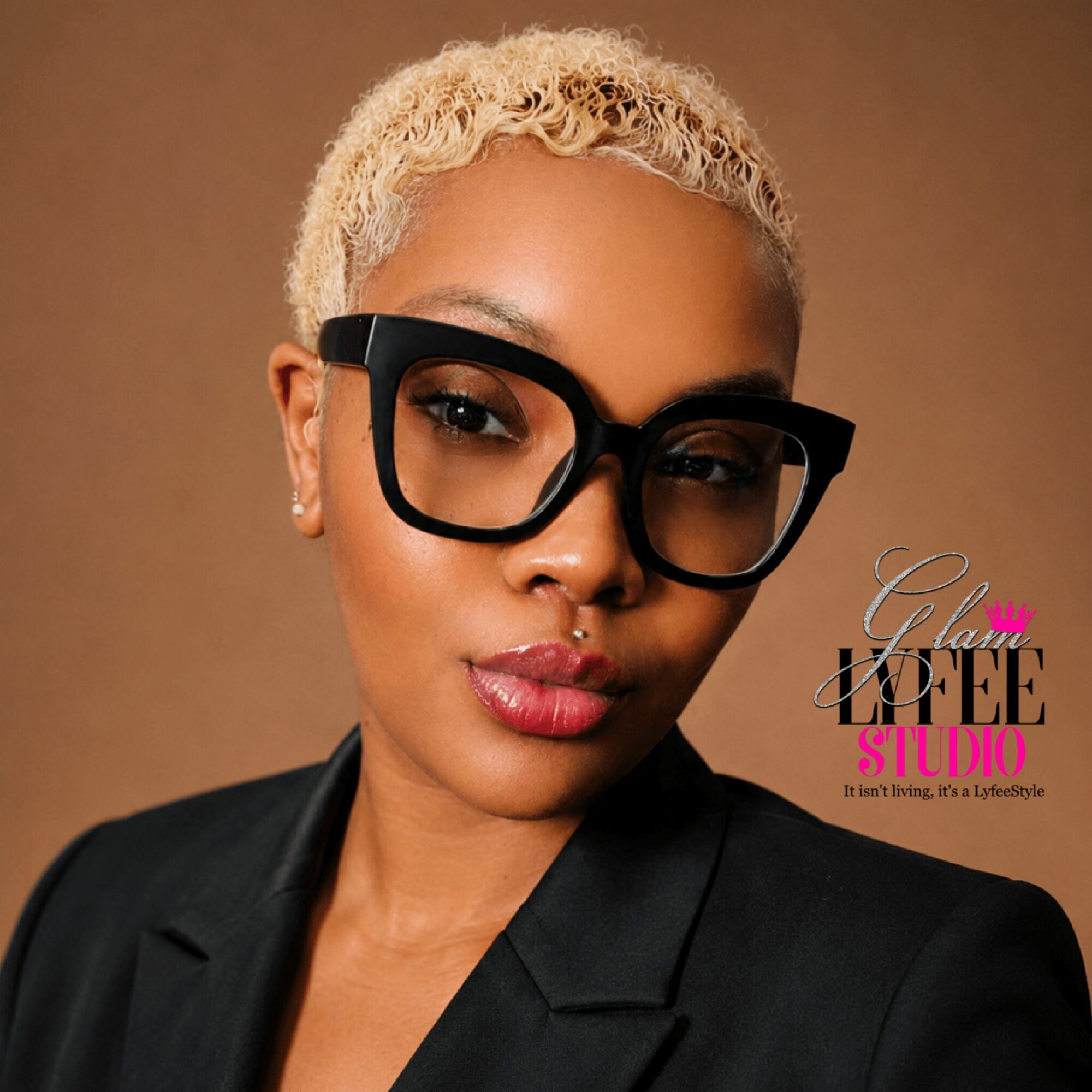 Woman with short blonde curly hair wearing large black glasses and a black blazer, looking at the camera.