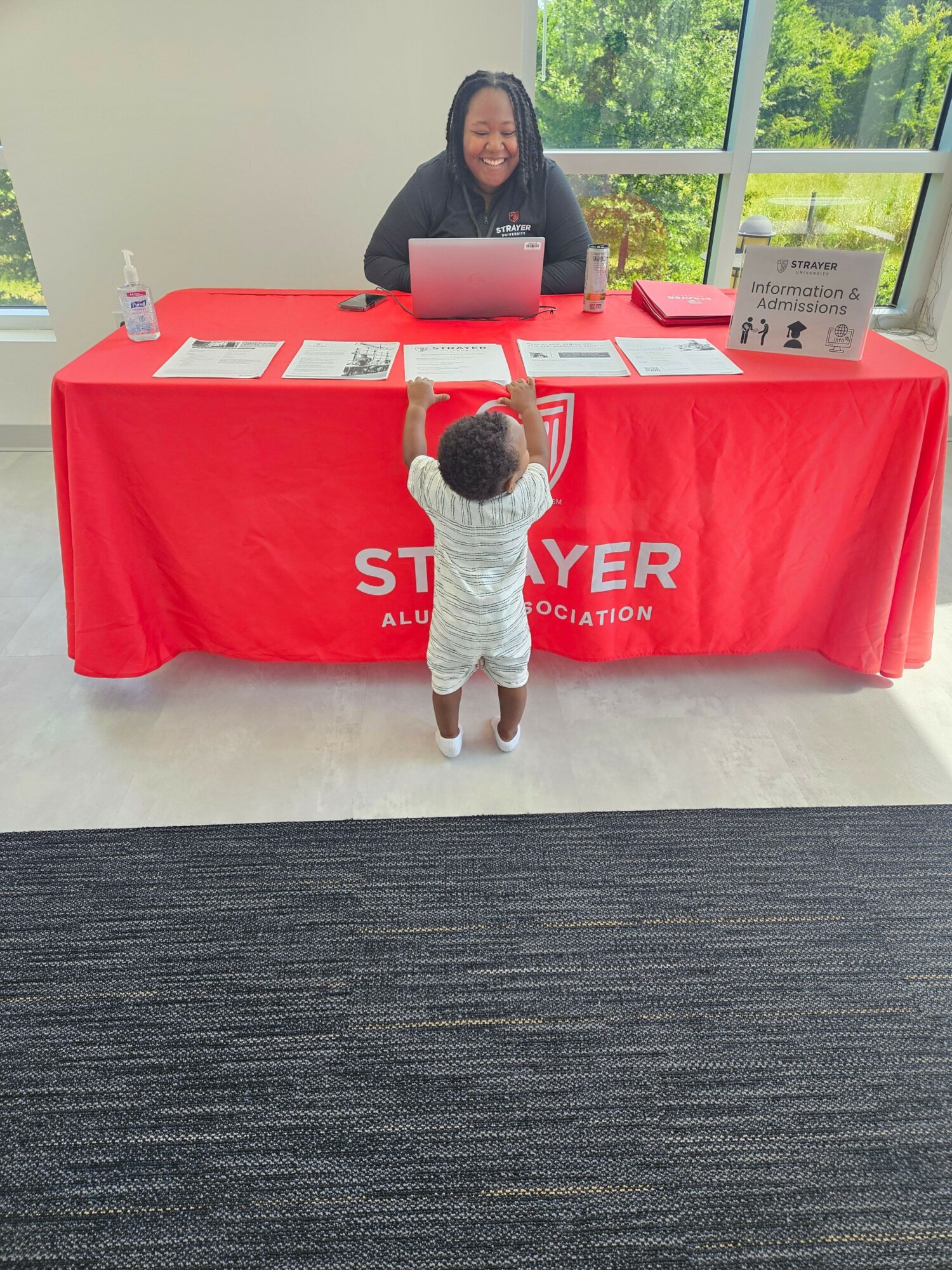 Child standing in front of a table with a red tablecloth, with a woman behind the table, in a room with large windows.