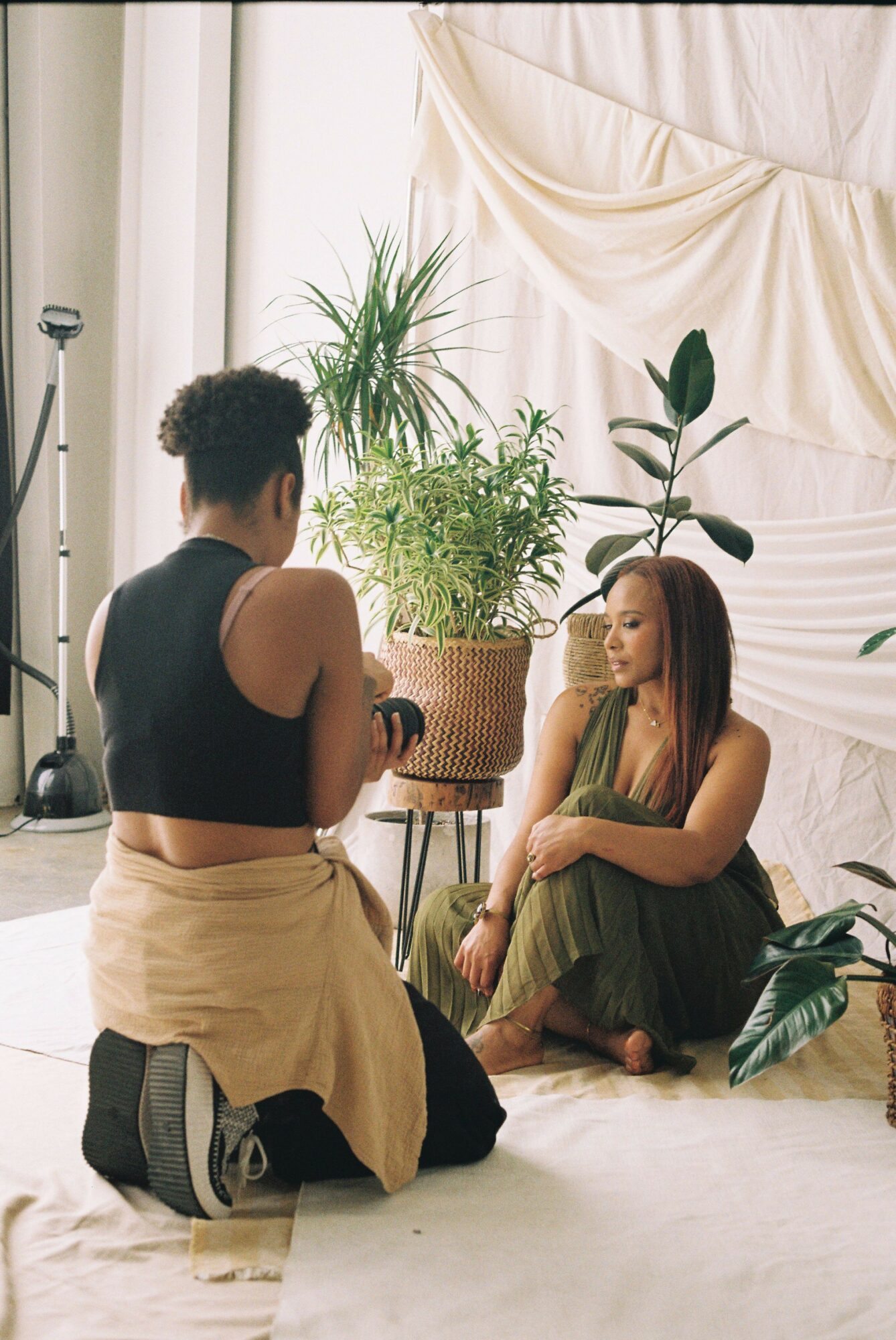 Two women sitting on the floor near plants and a window, one with curly hair and the other with straight hair, engaged in conversation.