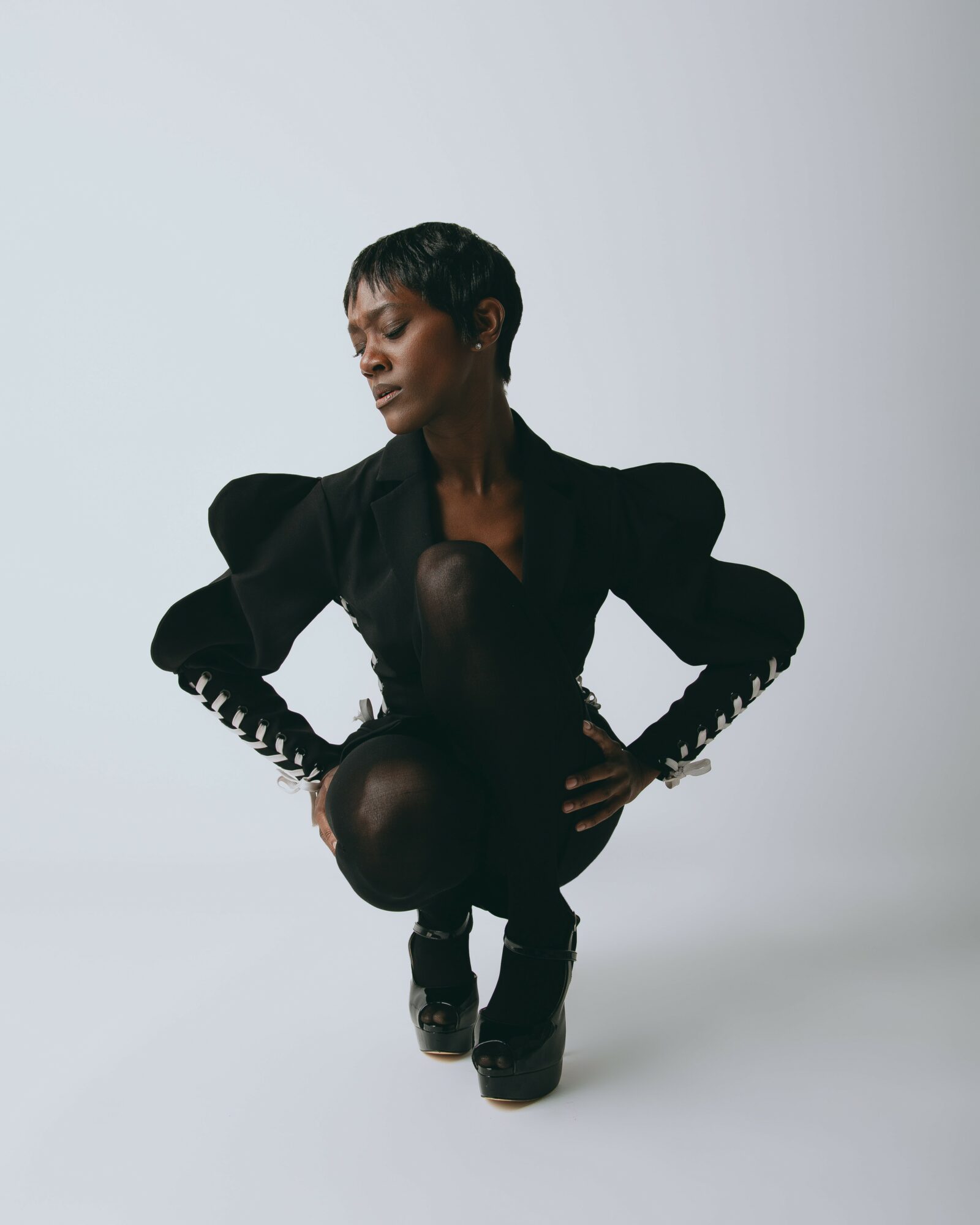 Woman crouching with hands on hips, wearing a black outfit with puffed sleeves, against a plain light background.