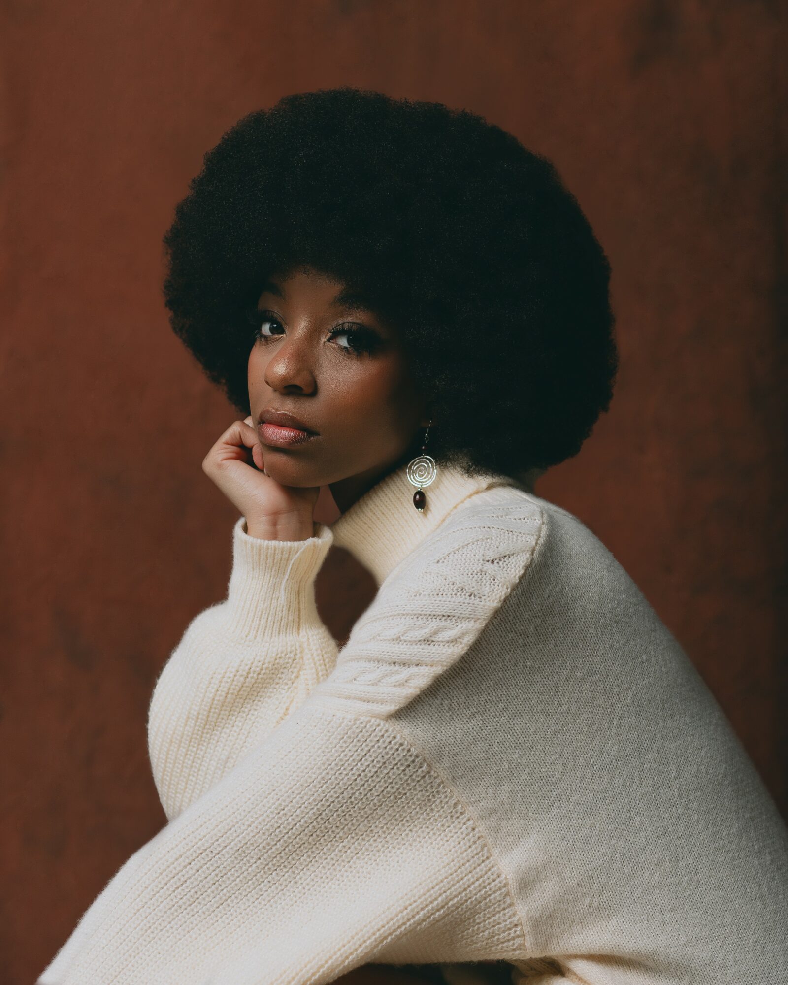 Portrait of a woman with a large afro hairstyle, wearing a cream sweater and earrings, resting her chin on her hand.