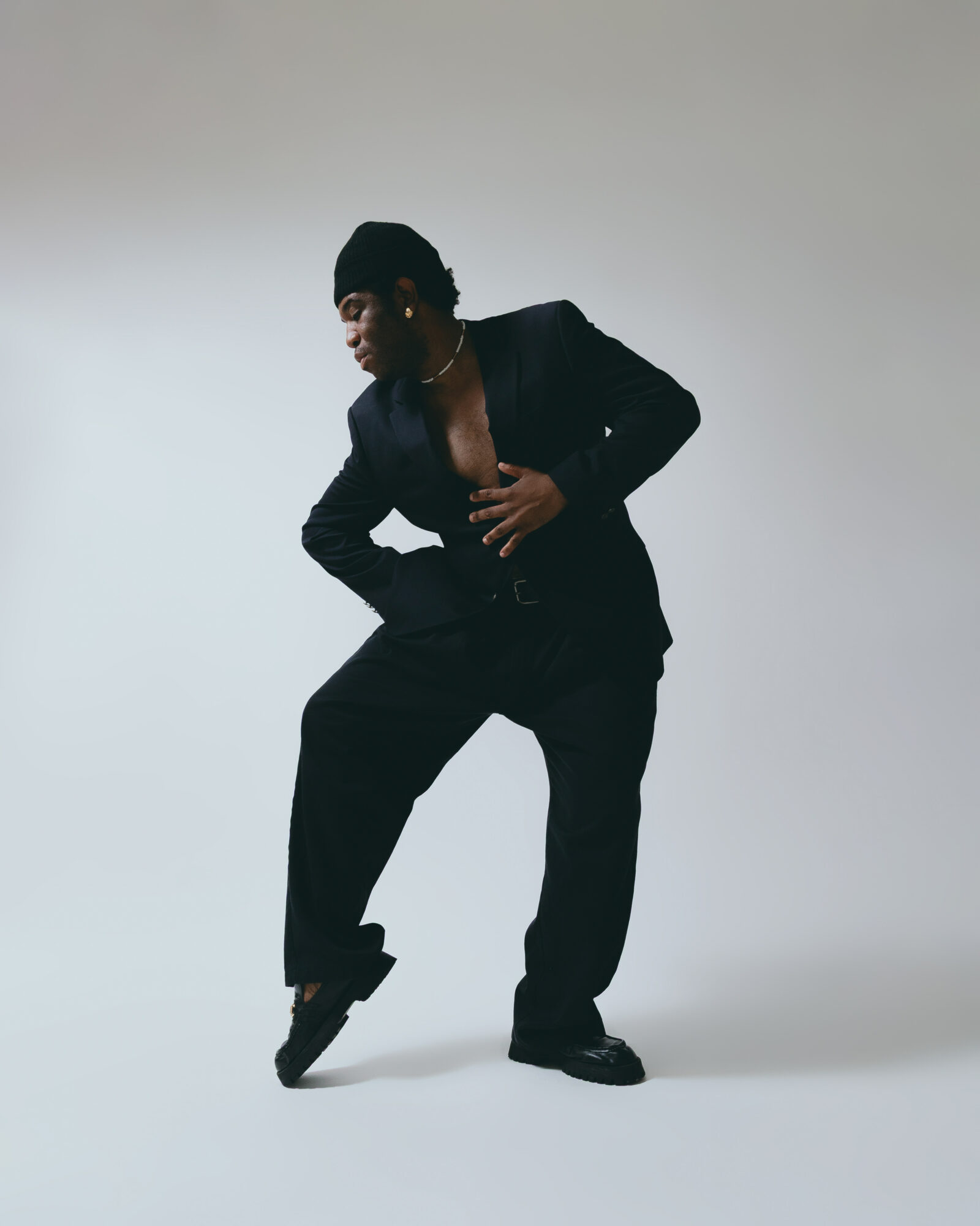 Person in black suit poses with one hand on stomach and the other on hip, against a plain background.