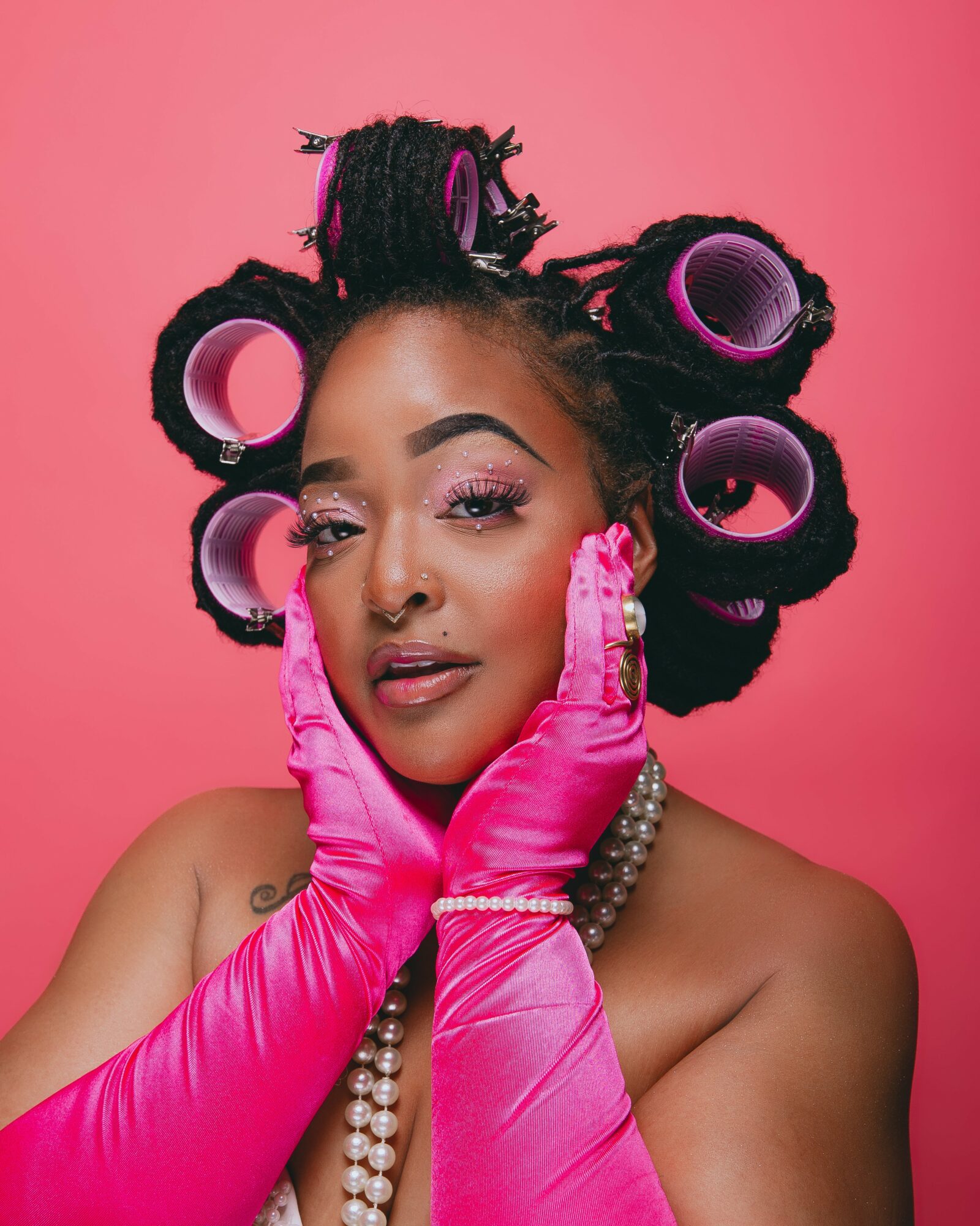Woman with styled hair in large rollers, pink gloves, and pearl necklace against pink background.