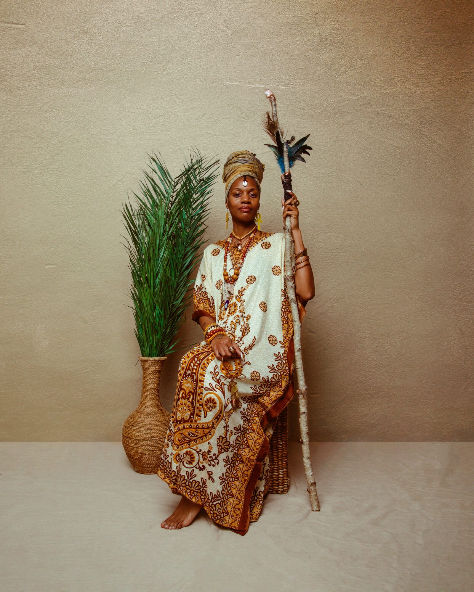 Person dressed in traditional attire holding a staff, standing next to a large plant in a woven basket, against a plain background.