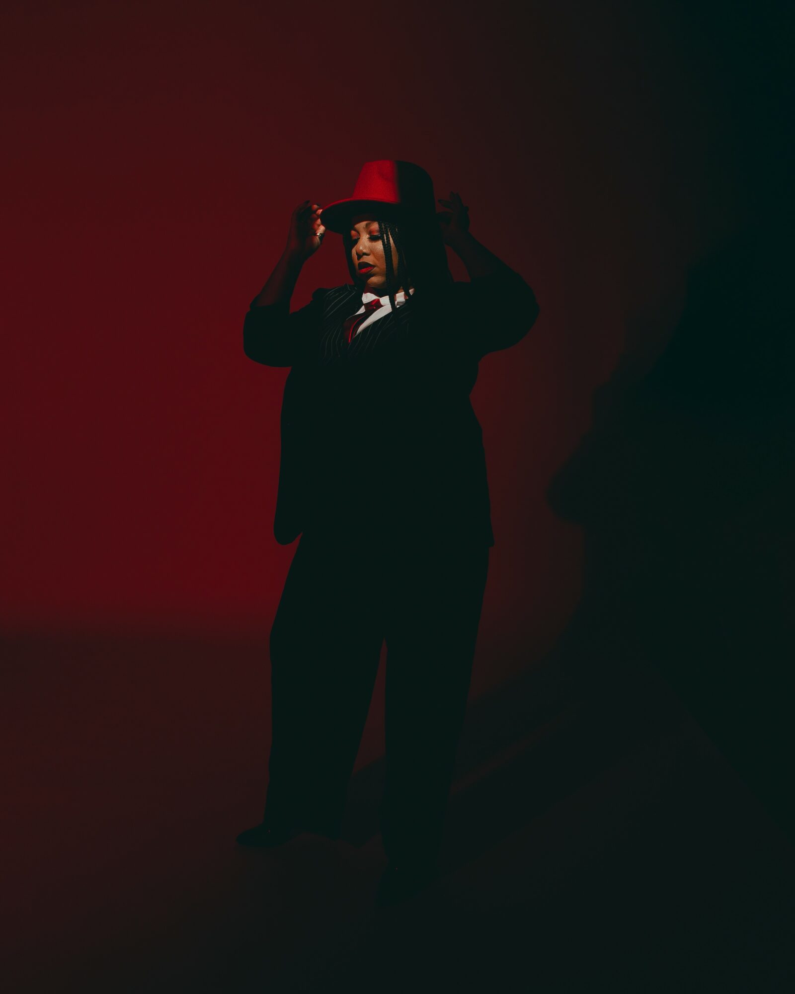 Person in a suit and red hat adjusting the hat, standing in a dark setting with red and black background.