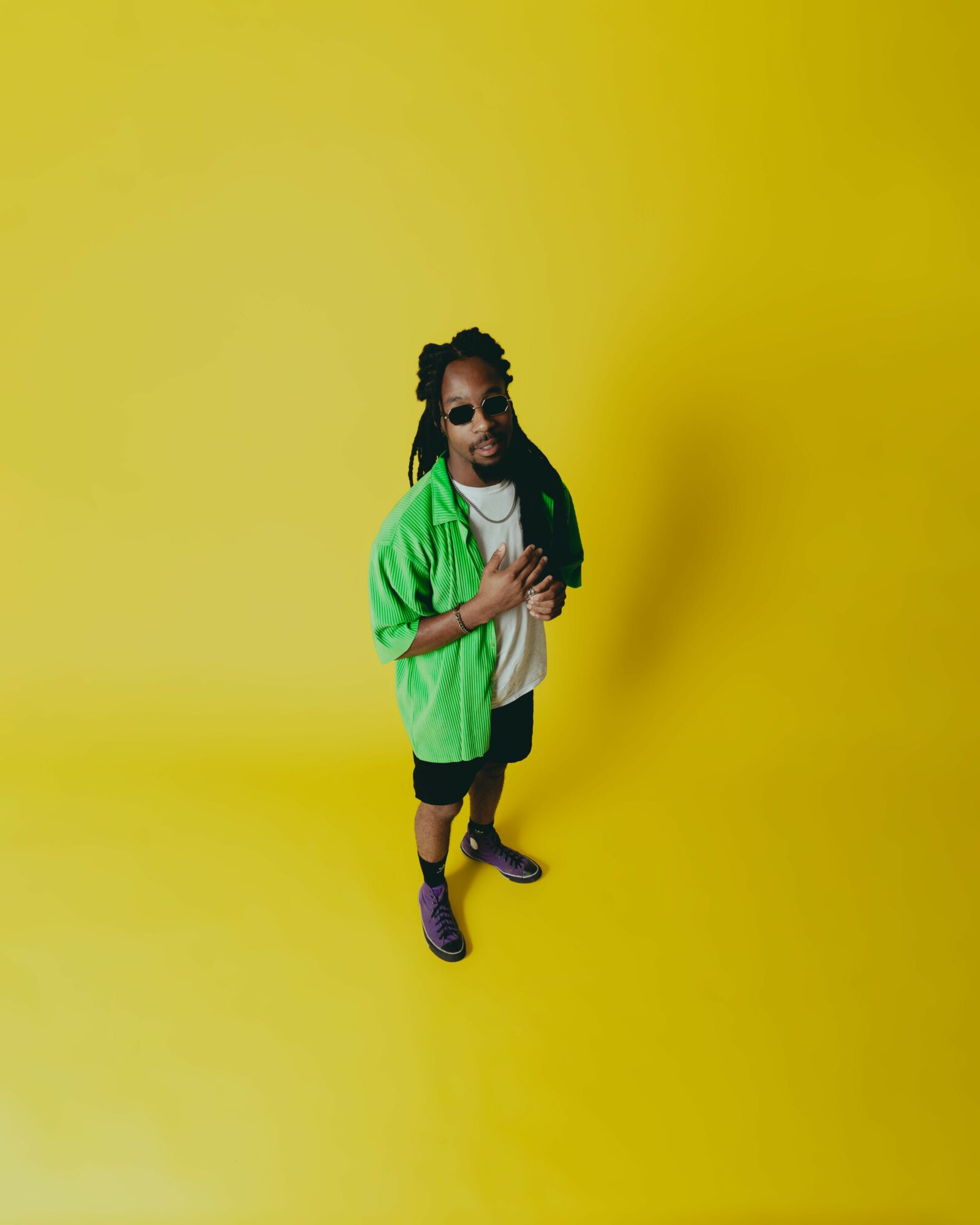 Person with long dreadlocks, sunglasses, green jacket, white shirt, black shorts, and purple shoes standing on yellow background.