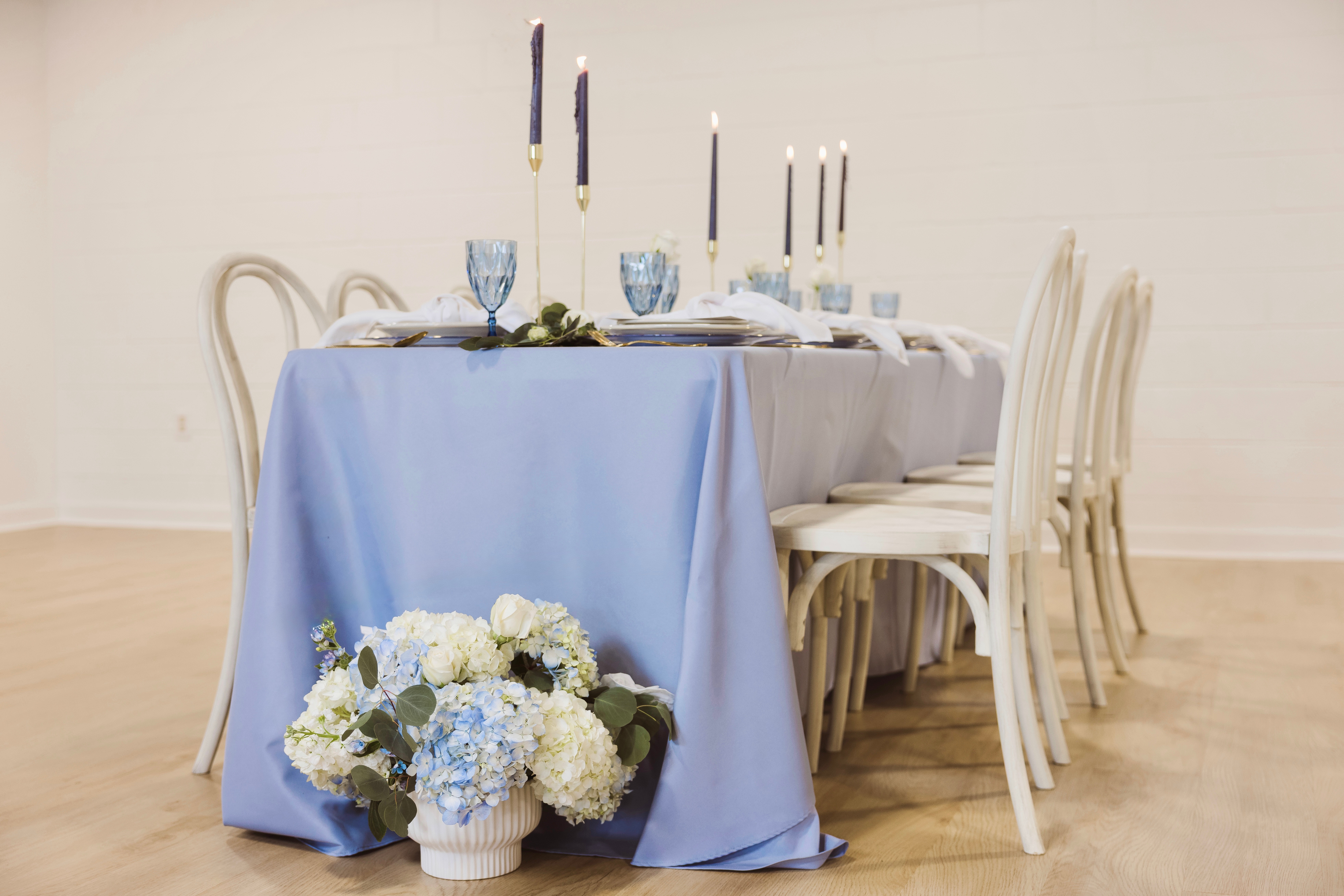 A dining table with a light blue tablecloth, set with candles, glasses, and plates, with a floral arrangement on the floor nearby.