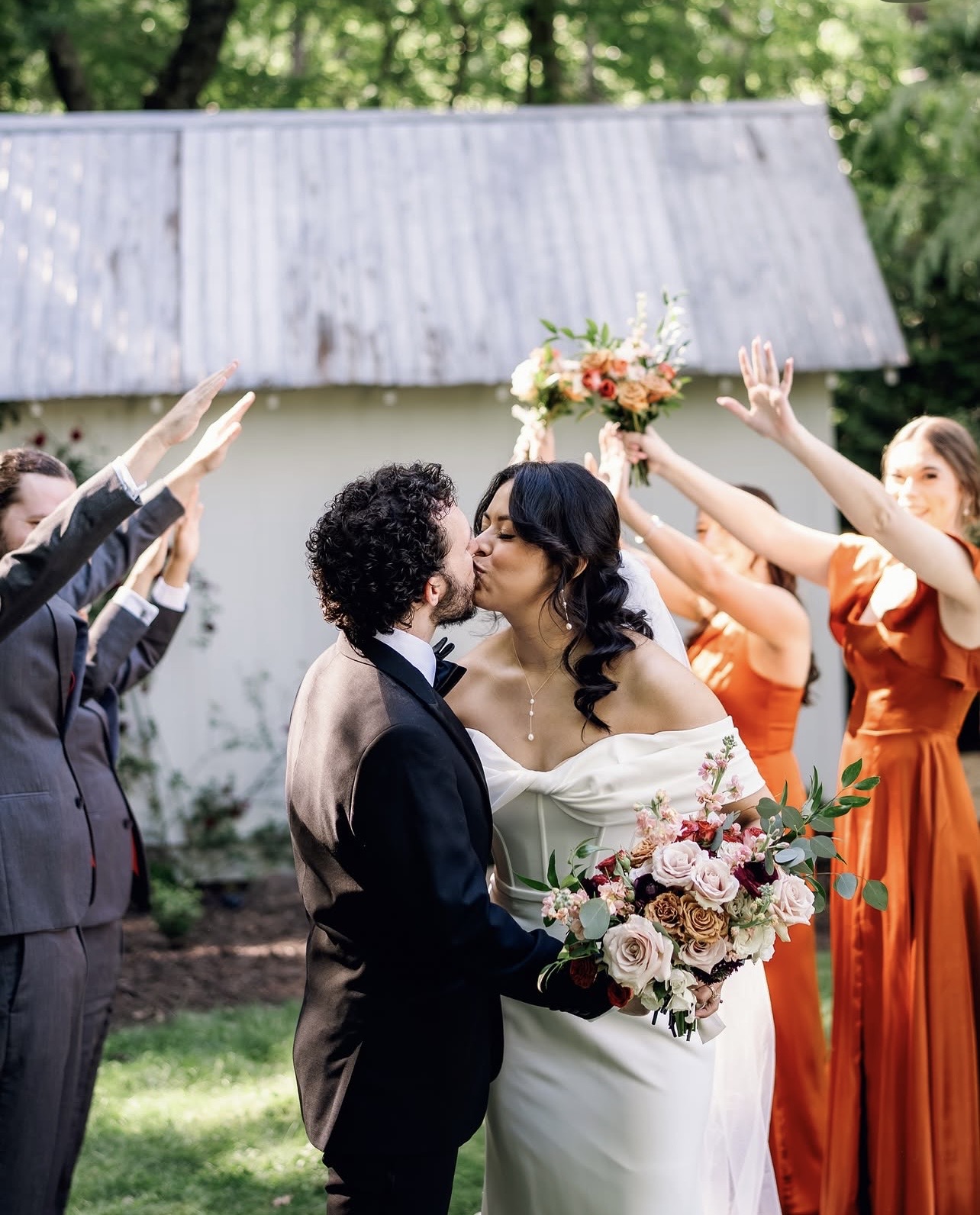 Couple kissing outdoors, surrounded by friends celebrating, some raising hands, with a rustic building in background.