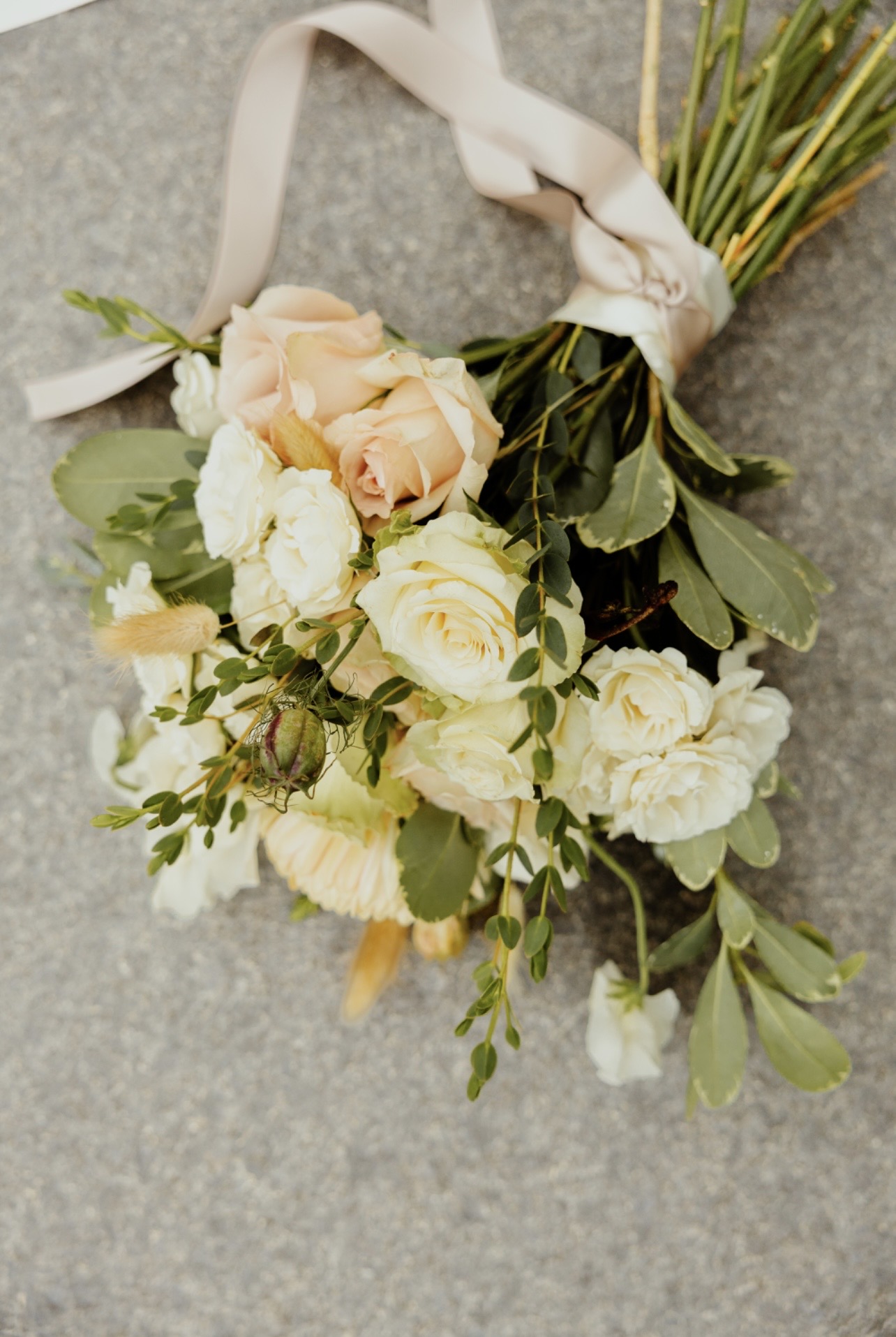 Bouquet of cream and peach roses with green leaves and a ribbon, placed on a gray surface.