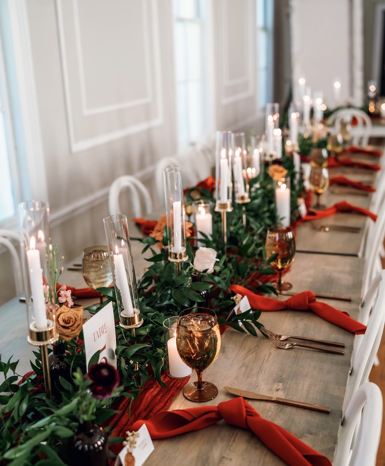 Long dining table decorated with candles, flowers, and red napkins, set for a formal event in a bright room.
