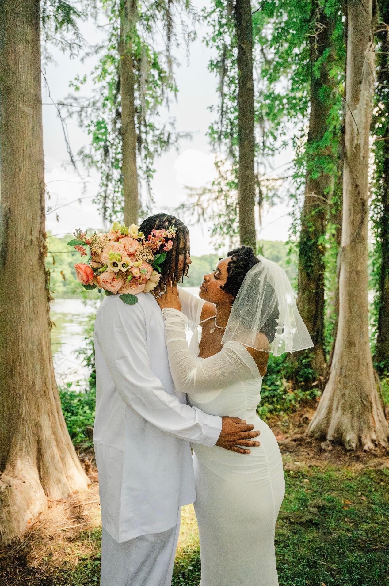 A couple in wedding attire stands close in a forest, holding a bouquet of flowers, with trees and sunlight in the background.