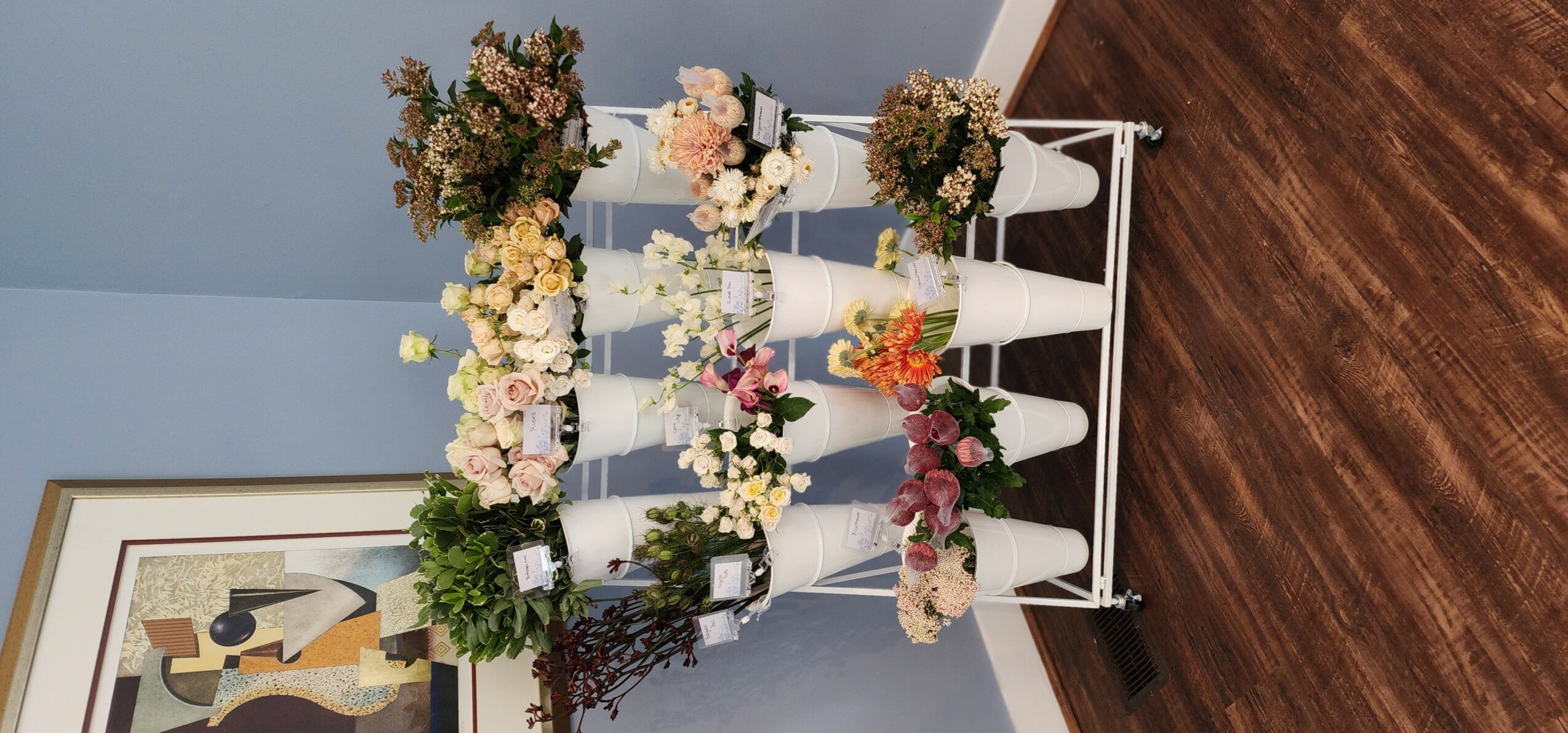 White wooden chair with multiple flower arrangements on a blue wall background, next to a framed artwork and wooden floor.