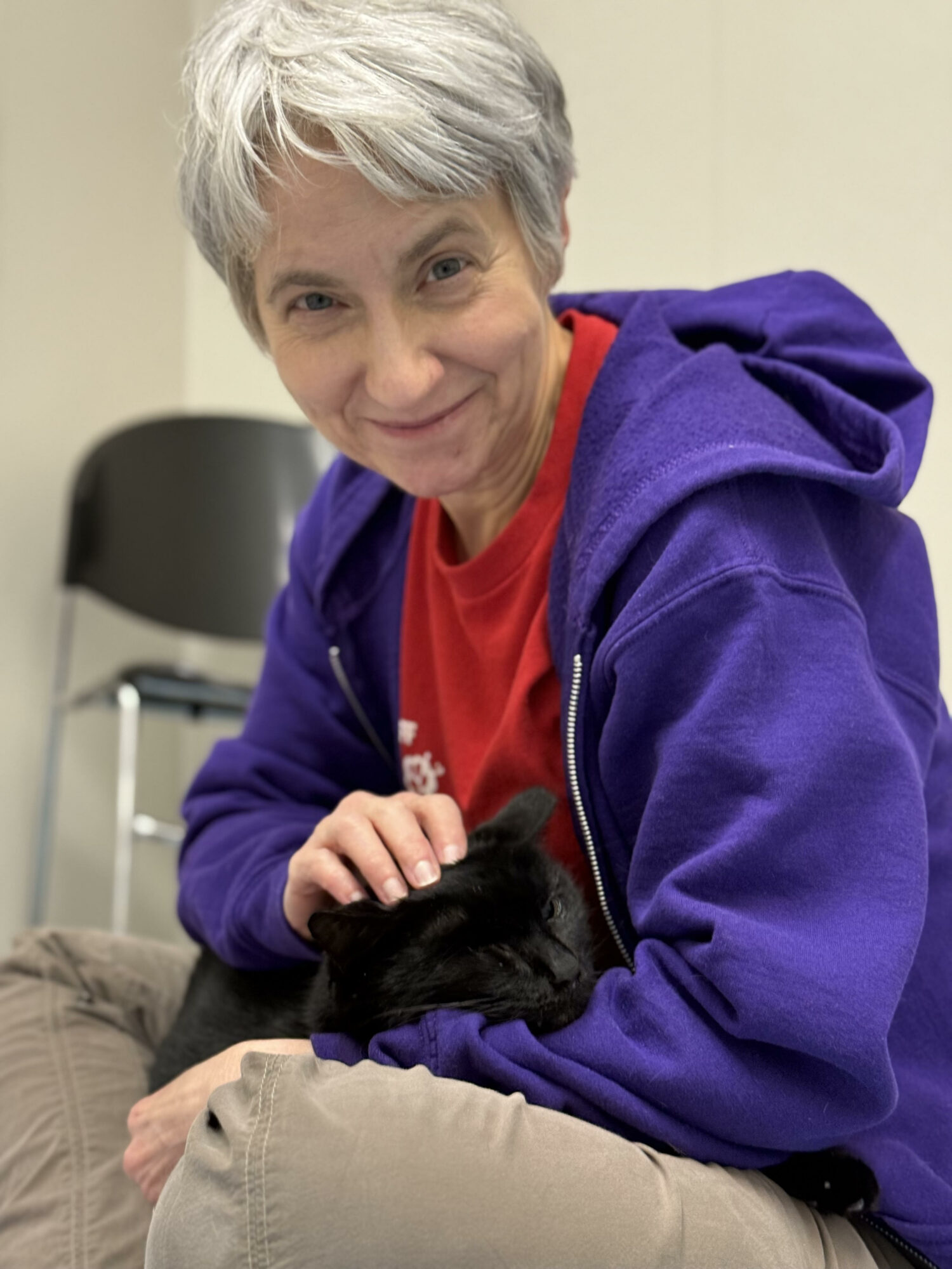 Woman with short gray hair smiling, wearing a purple hoodie and red shirt, holding a black cat.