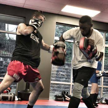 Two people sparring in a gym, wearing boxing gloves and athletic clothing, with windows and gym equipment in the background.