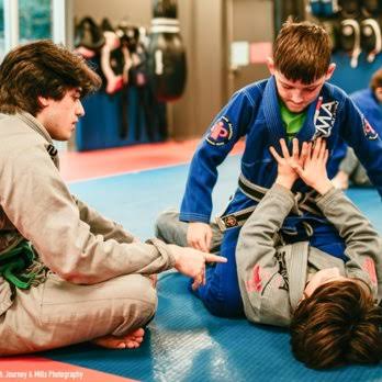 Two children practicing Brazilian Jiu-Jitsu with an instructor in a gym, one child on the ground and the other standing, both wearing gis.