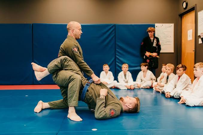 Martial arts instructor demonstrates a technique to children sitting on the floor in a dojo, with an instructor observing in the background.