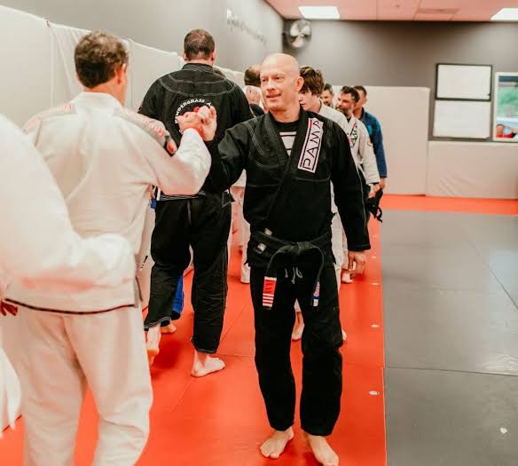 Two men in martial arts uniforms shake hands in a training room with other people in the background.