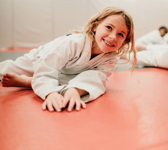 Young girl in a white martial arts uniform lying on a red mat, smiling at the camera.