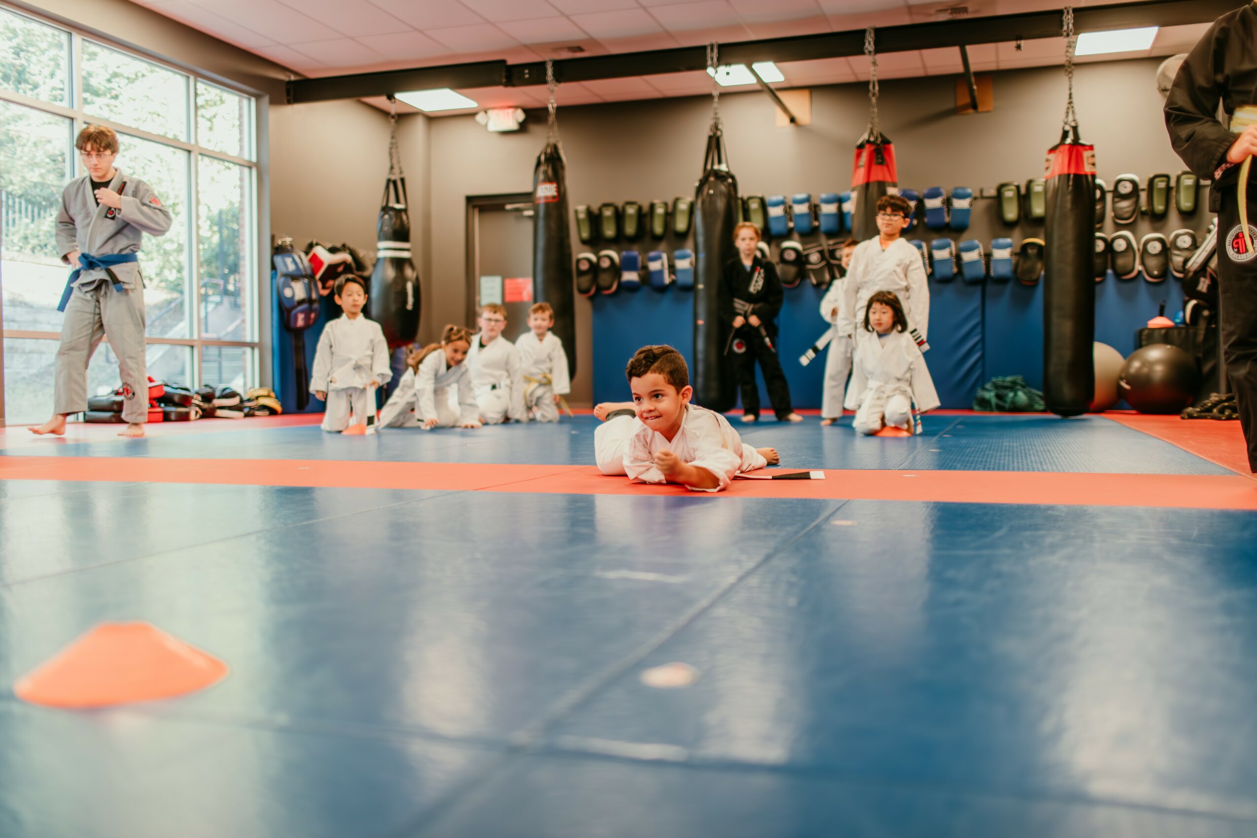 Children practicing martial arts on mats in a gym with punching bags and equipment, instructor supervising, large windows in background.