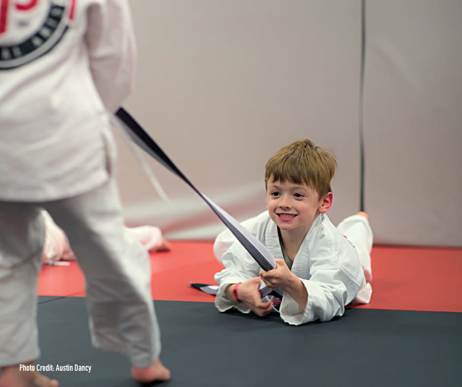 Child in a white martial arts uniform on a mat, smiling and holding a black belt, with another child partially visible.