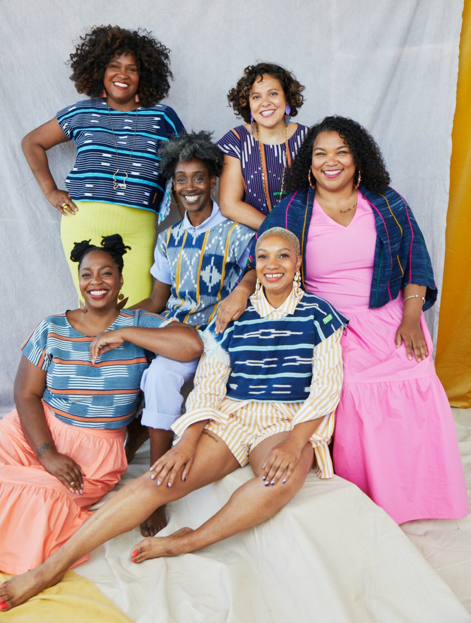 Six women with curly hair smiling, sitting and standing, wearing colorful casual clothing, against a light background.