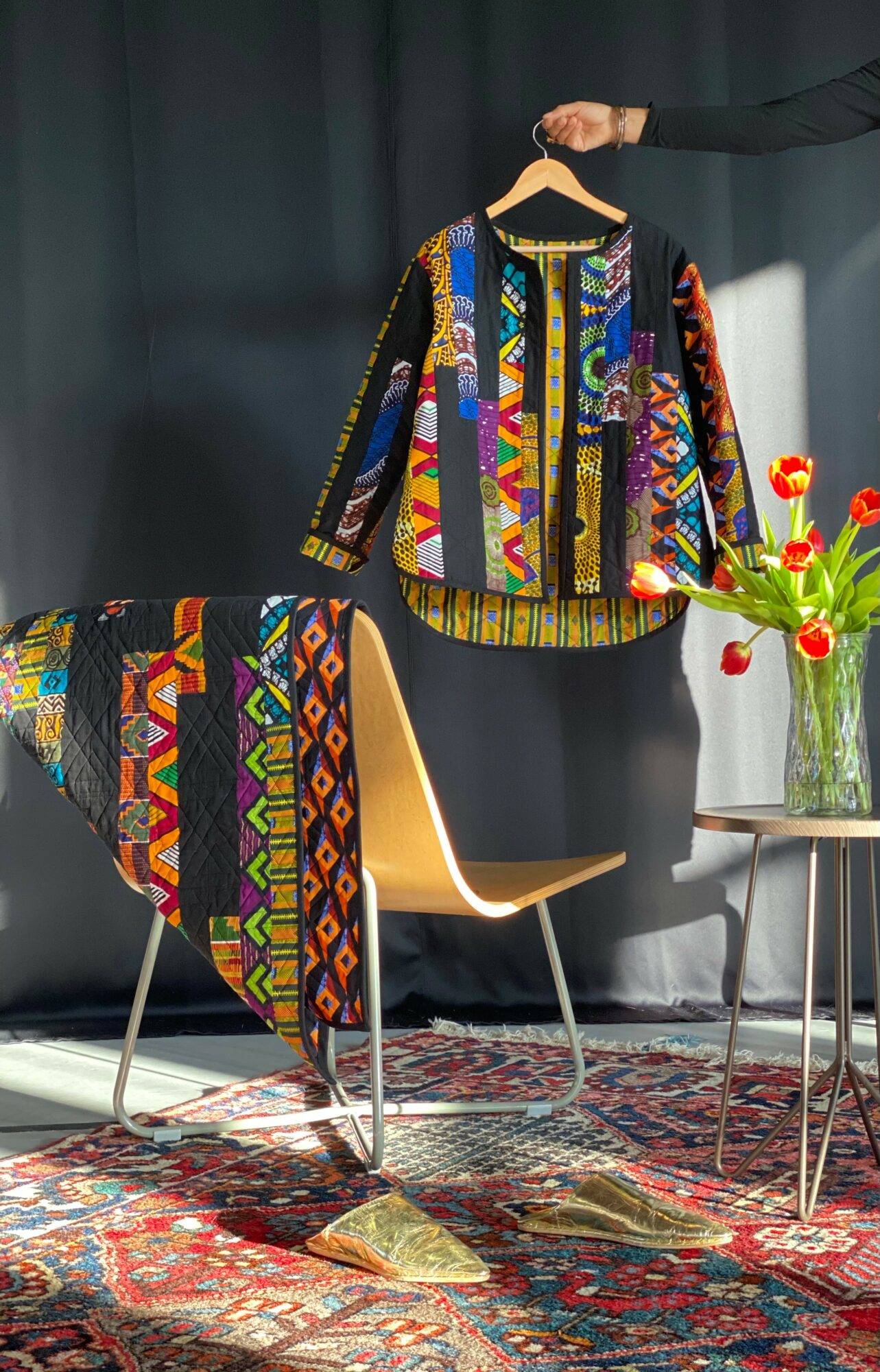 Colorful embroidered jacket hanging on a hanger, with a matching cloth draped over a chair, next to a vase of red tulips on a table.