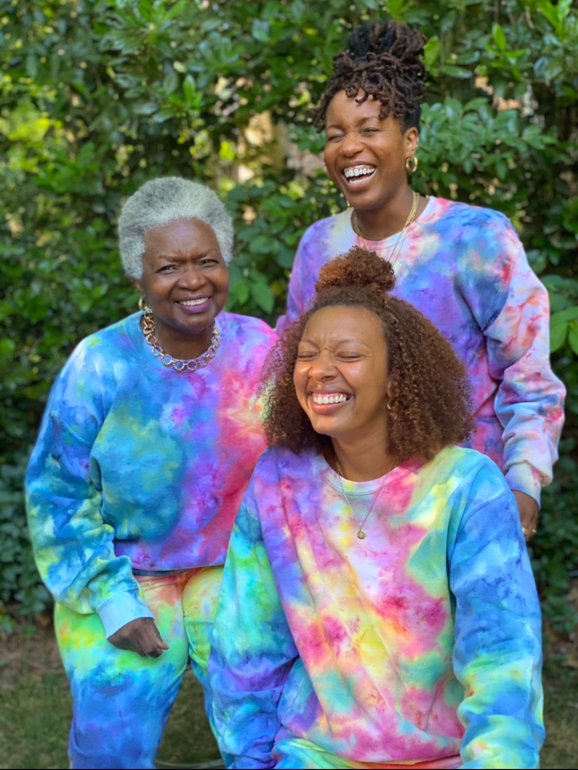 Three women smiling and laughing outdoors, wearing colorful tie-dye sweatshirts, with green foliage in the background.