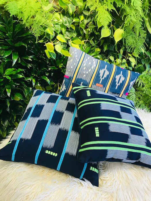 Three decorative pillows with blue, green, and black patterns are placed on a fluffy surface in front of green foliage.