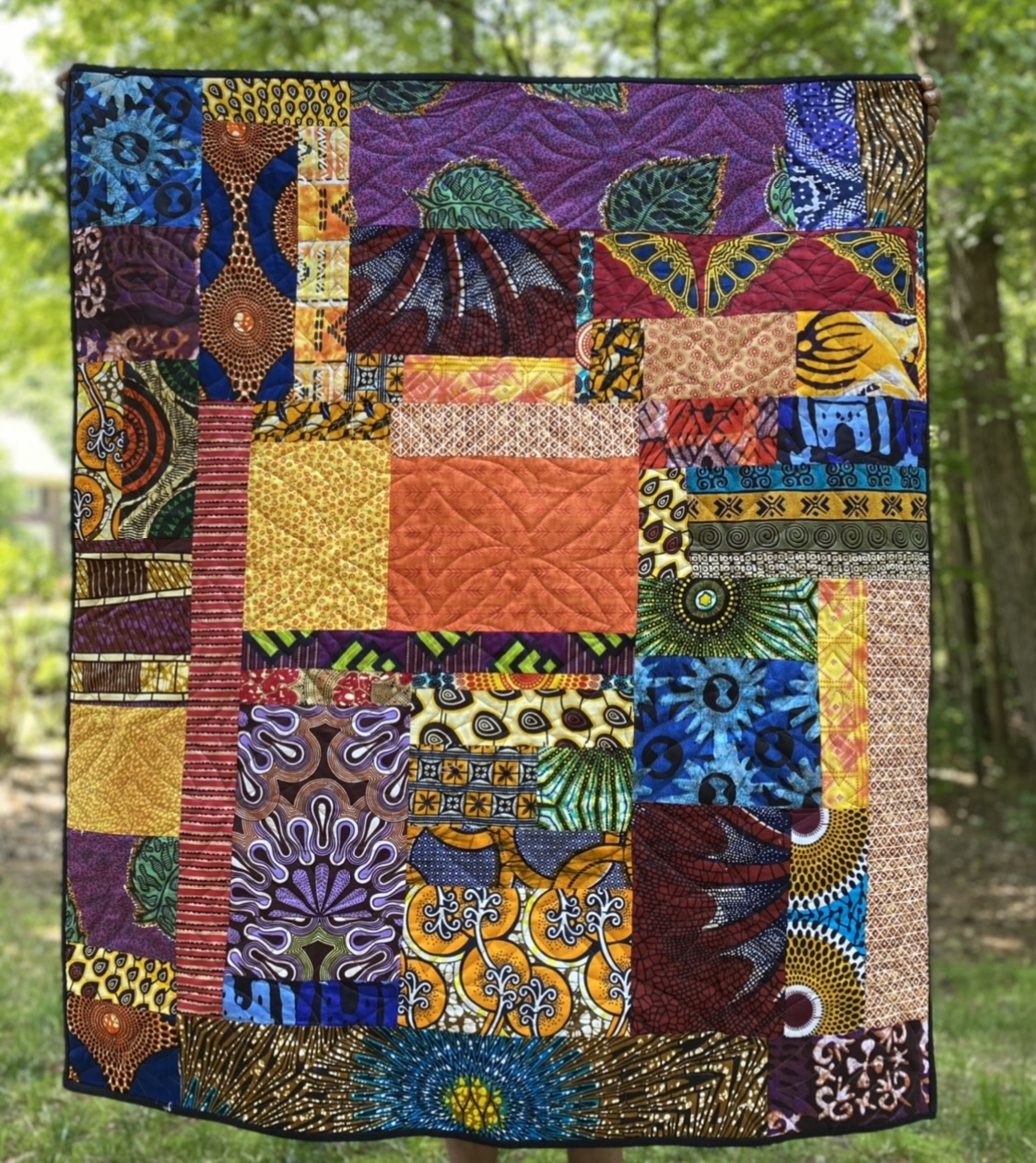 Colorful patchwork quilt with various patterns and colors, hanging outdoors with trees in the background.