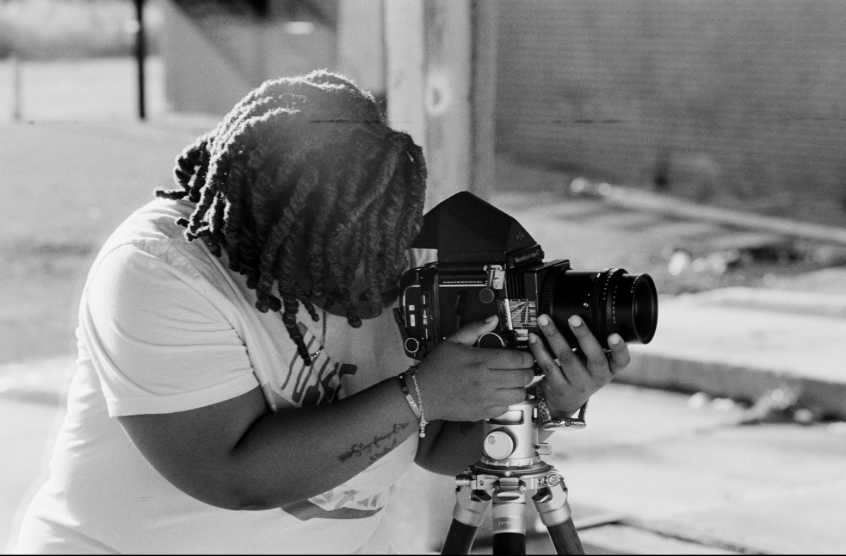 Person with dreadlocks looking through a camera on a tripod outdoors, in black and white.