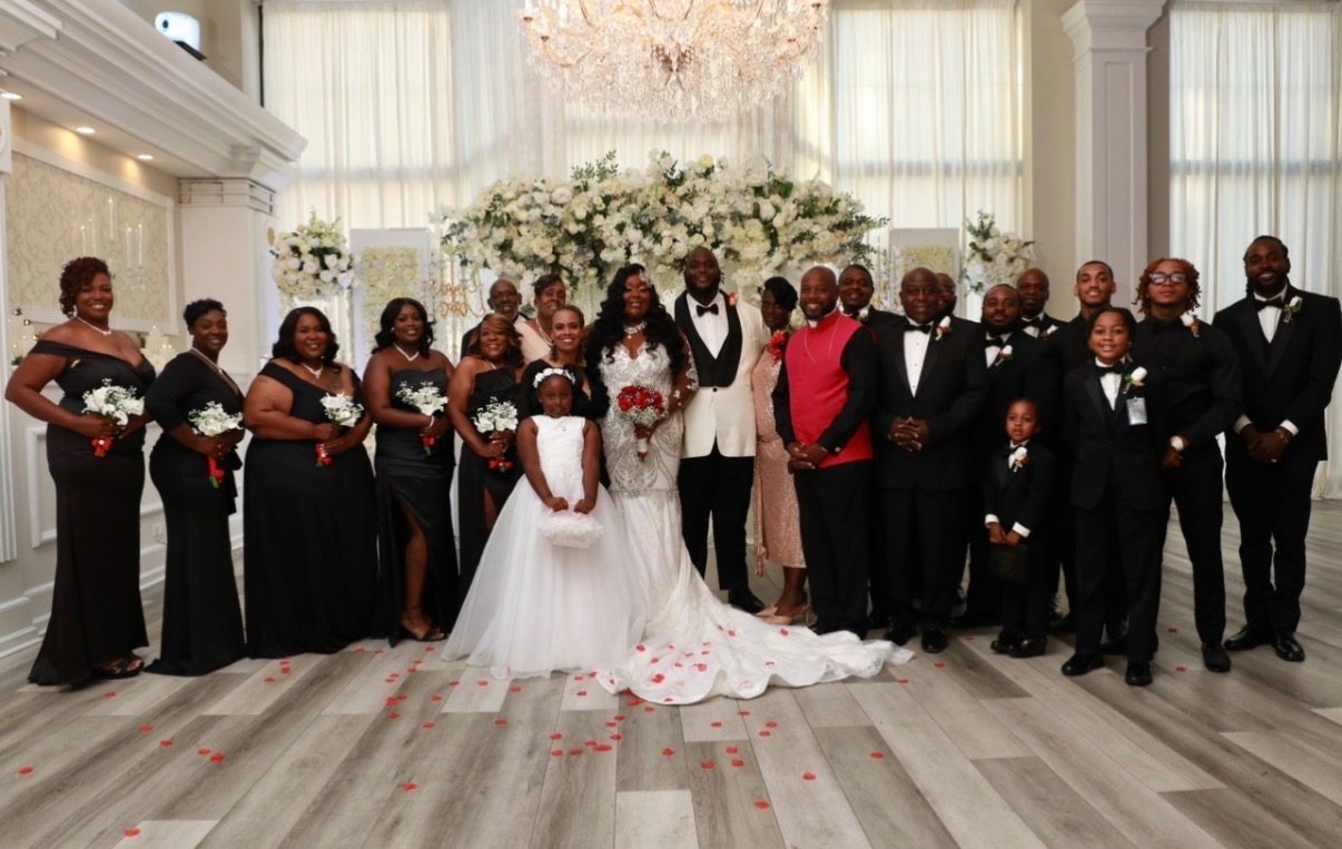 Group of people in formal attire at a wedding, with a bride and groom in the center, in a decorated hall.