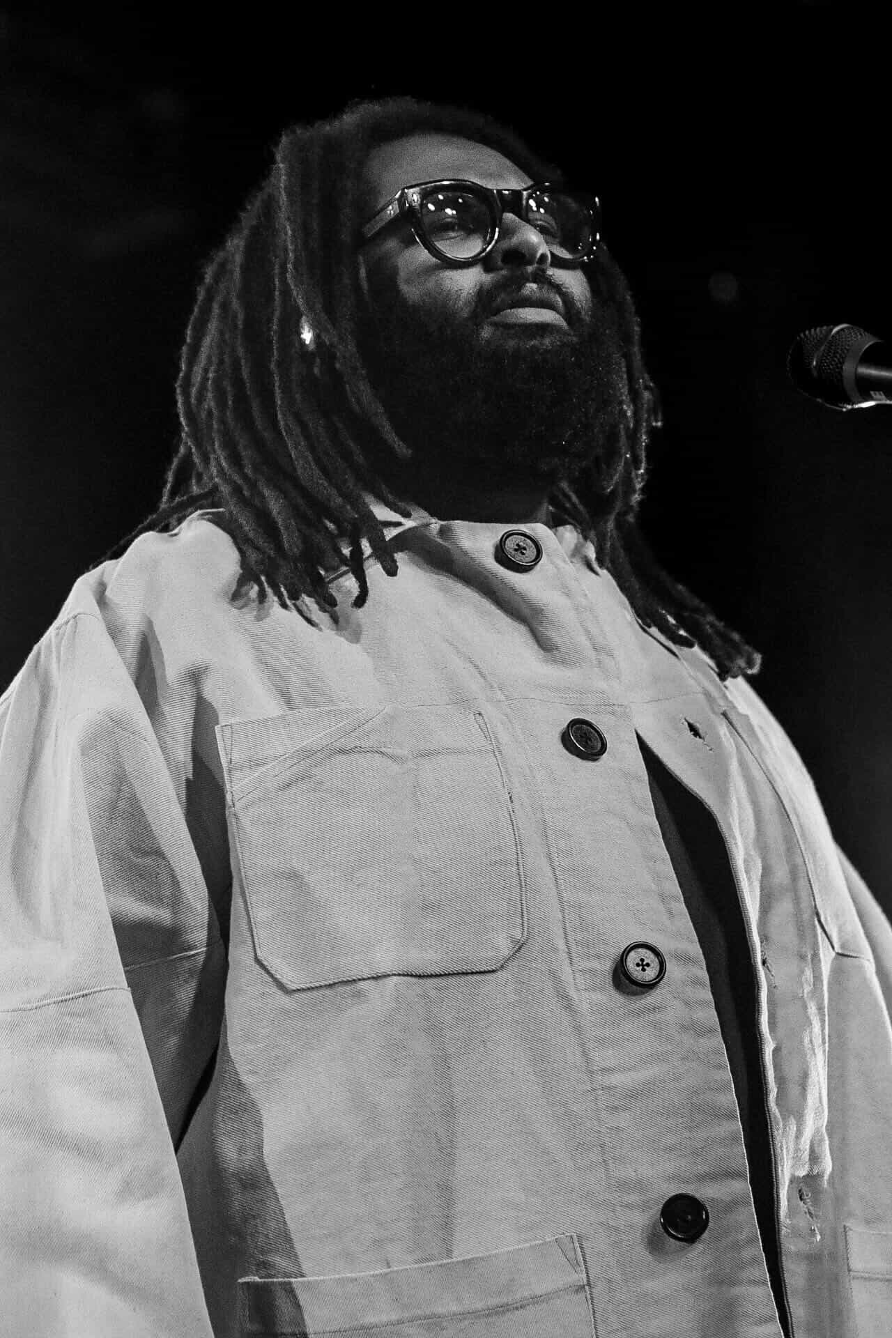 Person with glasses and dreadlocks speaking into a microphone, wearing a light-colored jacket, in black and white.
